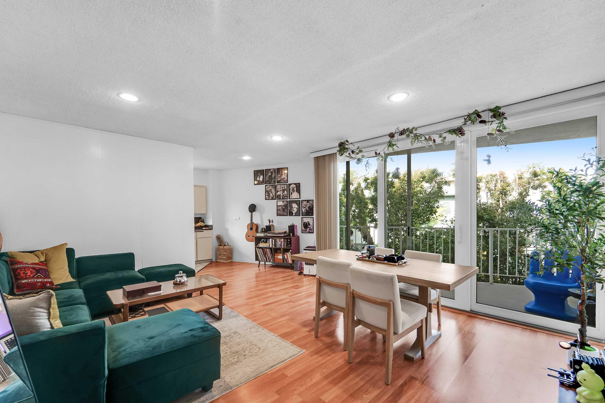 Living room featuring a green sectional sofa and wooden dining table. The space has large windows with greenery outside, and walls adorned with framed photographs. A guitar stands in the corner, and decorative plants enhance the welcoming atmosphere. Light floods the room, creating a cozy ambiance.
