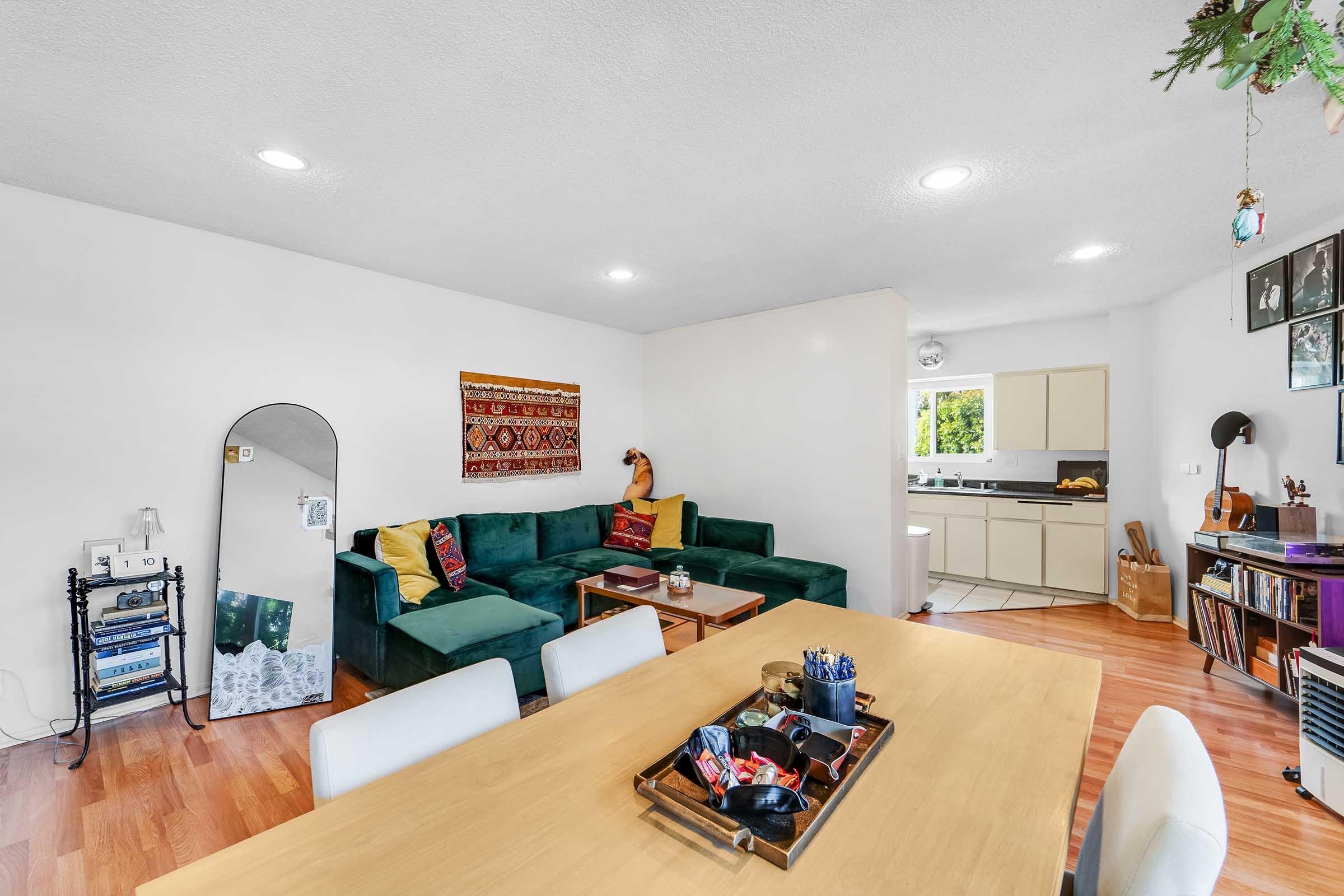 A cozy living area featuring a green sectional sofa adorned with colorful pillows, a wooden dining table with decorative items, and a floor mirror. In the background, there's a light kitchen with white cabinets and an adjacent window letting in natural light. The walls are decorated with art and plants.