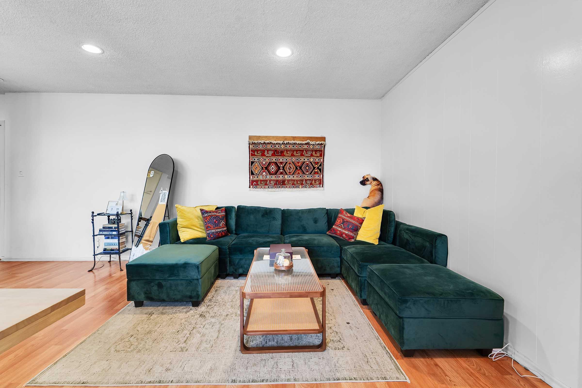 A cozy living room featuring a dark green sectional sofa adorned with yellow pillows and decorative cushions. There's a wooden coffee table in front, and a large wall mirror beside it. A patterned wall hanging adds warmth to the space, along with a plush area rug on the hardwood floor.