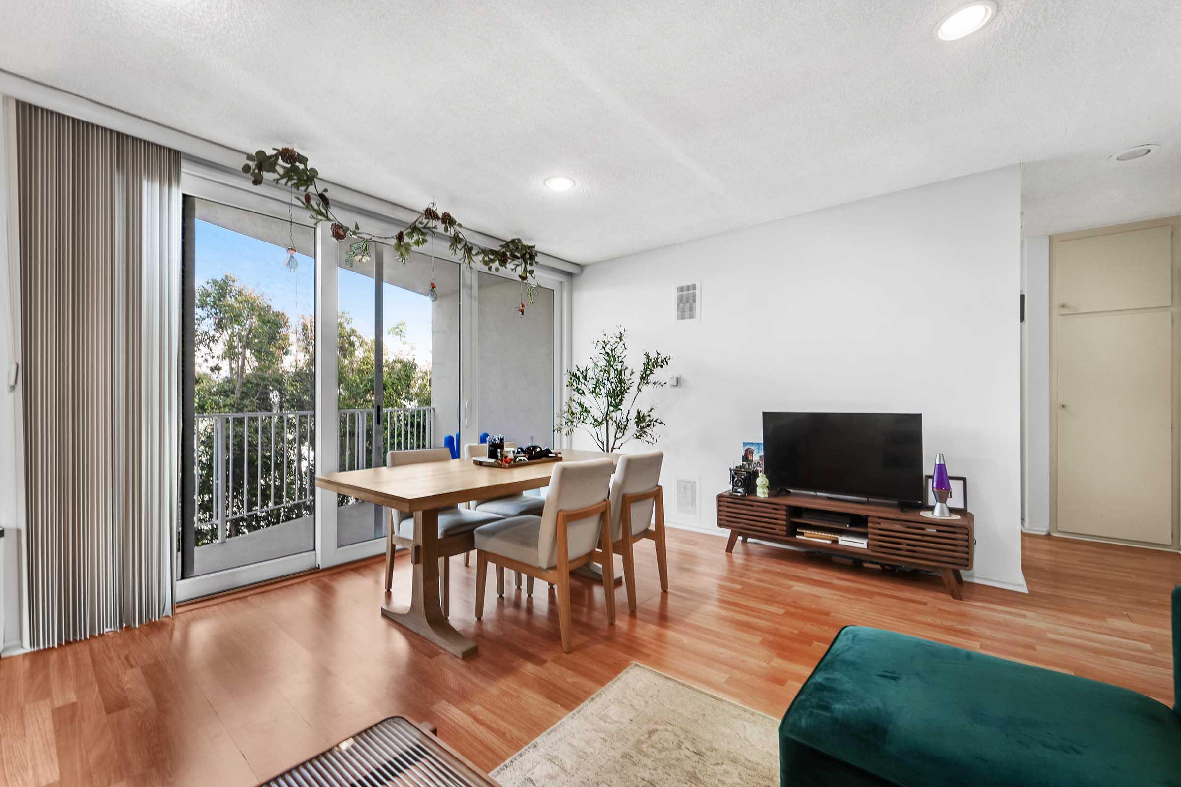 A bright, modern living area featuring a wooden dining table with chairs, a television on a mid-century style stand, and a green ottoman. Large glass doors lead to a balcony with greenery outside, while natural light fills the room, enhancing the clean, minimalist decor.