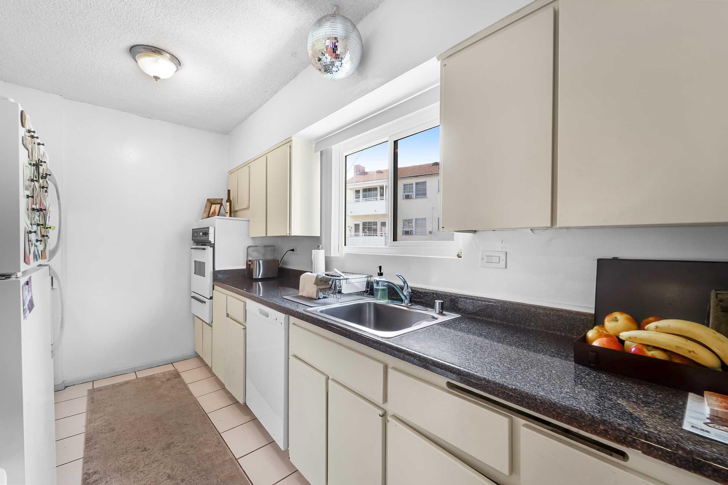 Bright kitchen featuring beige cabinets, a black countertop, and a window letting in natural light. There is a sink with dish soap next to a fruit bowl containing bananas and other fruits. A fridge adorned with magnets is visible, along with an oven and an area rug on the tiled floor.