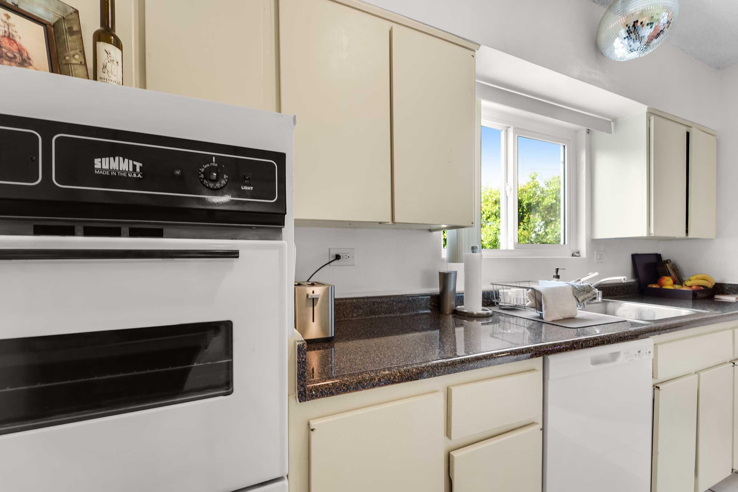 A modern kitchen featuring a white refrigerator and a stove. The countertop is dark and has a dish rack, coffee maker, and some fruit. Sunlight streams through a window with white cabinetry above. A disco ball hangs from the ceiling, adding a unique touch to the space.