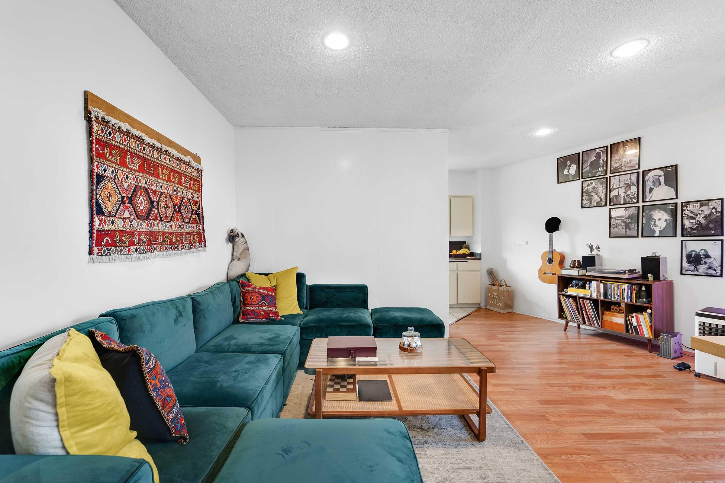 A cozy living room featuring a large teal sectional couch with colorful pillows, a wooden coffee table, and decorative items. The wall is adorned with a vibrant tapestry and framed photographs. In the background, a guitar rests against a shelf filled with books and other items, creating a warm and inviting atmosphere.