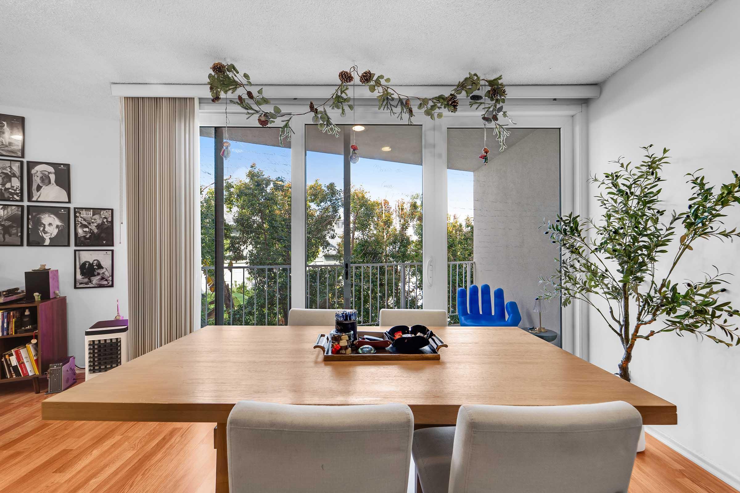 A modern dining area featuring a wooden table with a tray of items on it. There are two light-colored upholstered chairs. In the background, large sliding glass doors open to a balcony with greenery outside. Wall decor includes black and white photos. Natural light fills the space, creating a warm atmosphere.