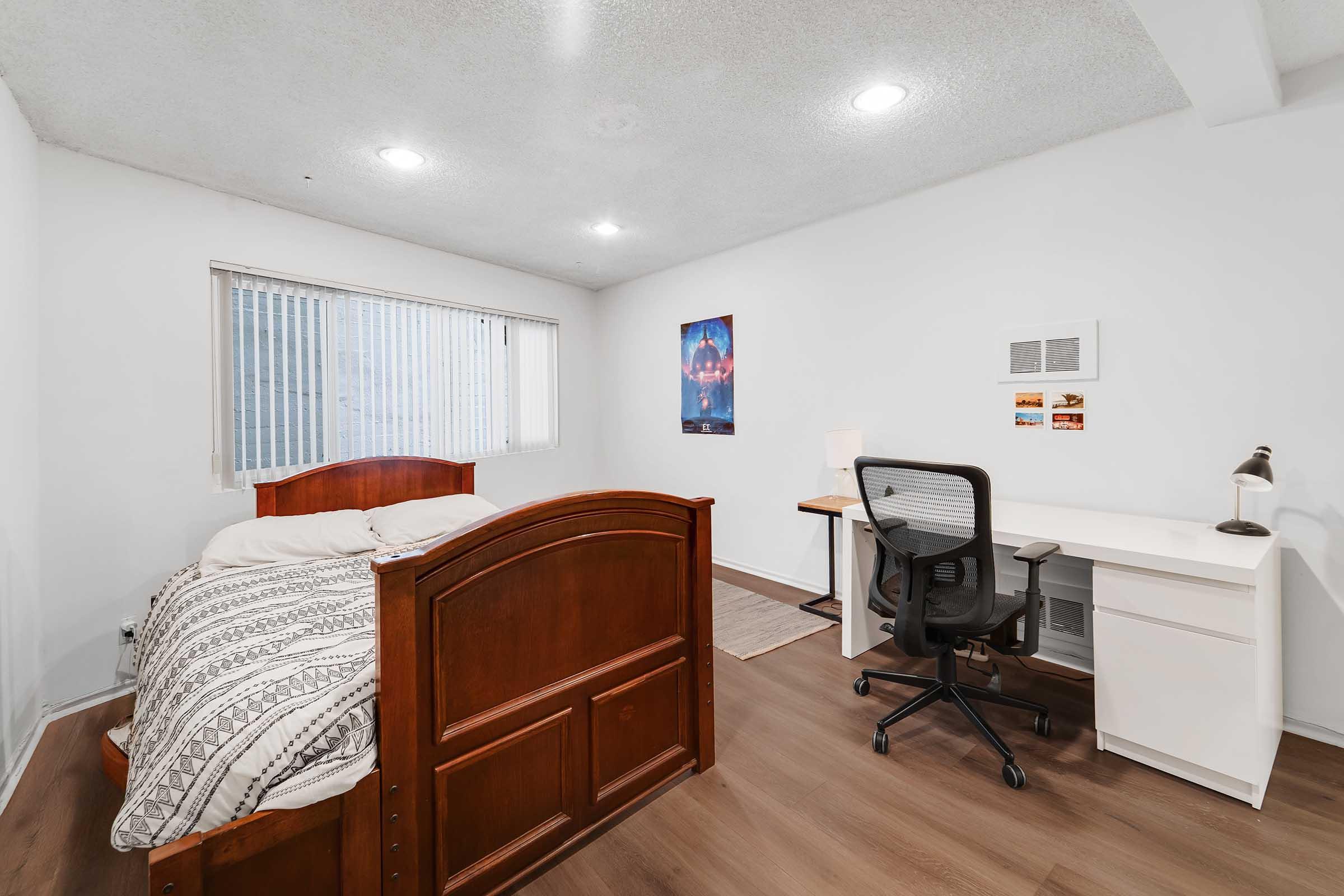 A cozy bedroom featuring a wooden bed with a patterned comforter, a desk with a chair, and a small lamp. The room is well-lit with natural light coming through a window with blinds. There is a framed poster on the wall and light-colored flooring, creating a spacious and inviting atmosphere.