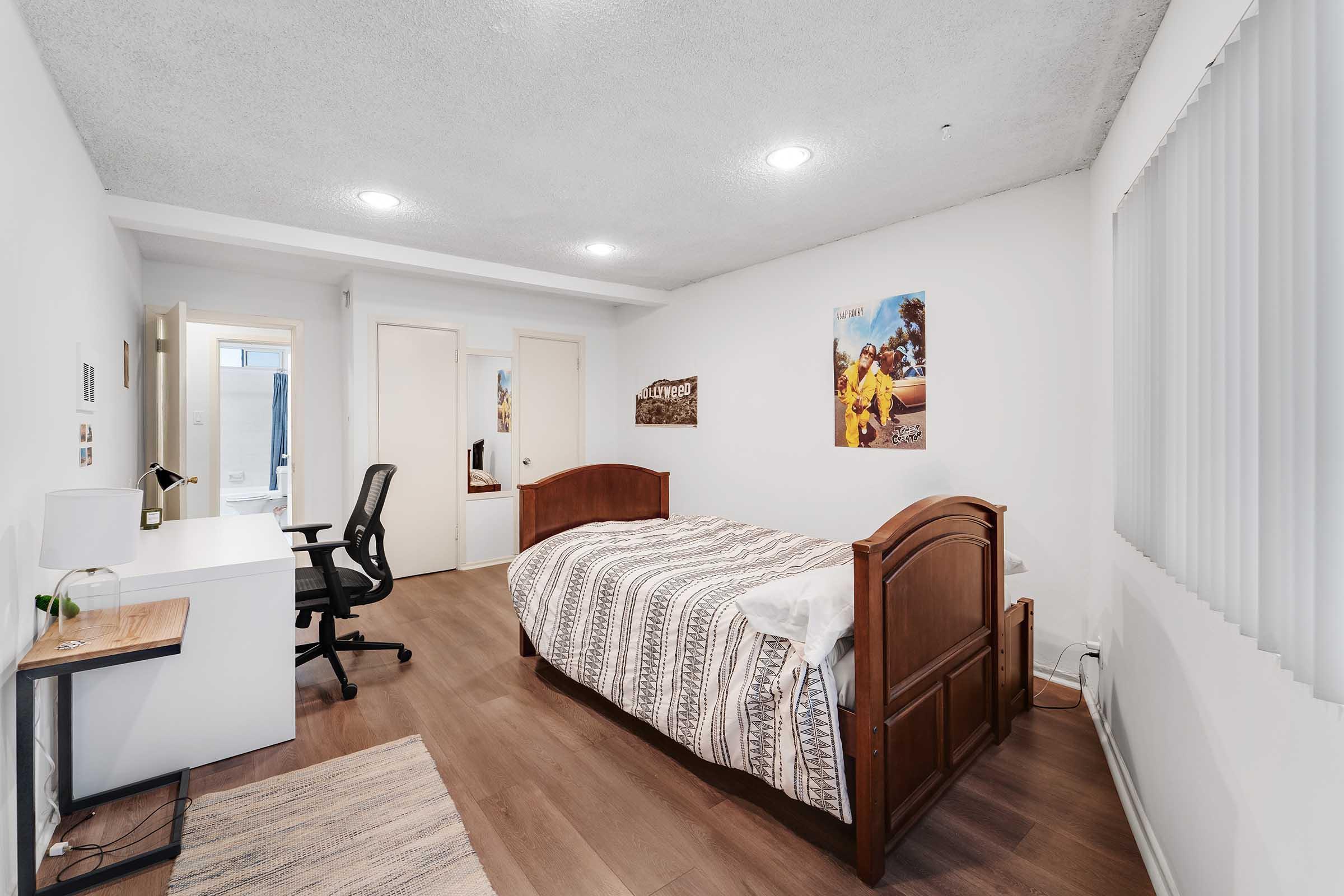 Cozy bedroom featuring a wooden bed with a patterned blanket, a white desk with a chair, and a large window with vertical blinds. The walls are painted white, and there are two posters on the walls. The floor is hardwood, creating a warm and inviting atmosphere.