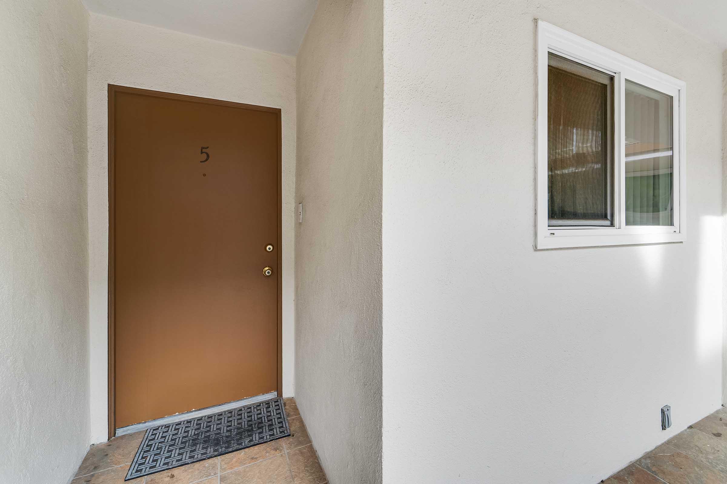 A simple entrance with a brown door labeled "5" and a small window to the right. The walls are light-colored and the floor features tiled flooring. A black doormat is placed in front of the door.