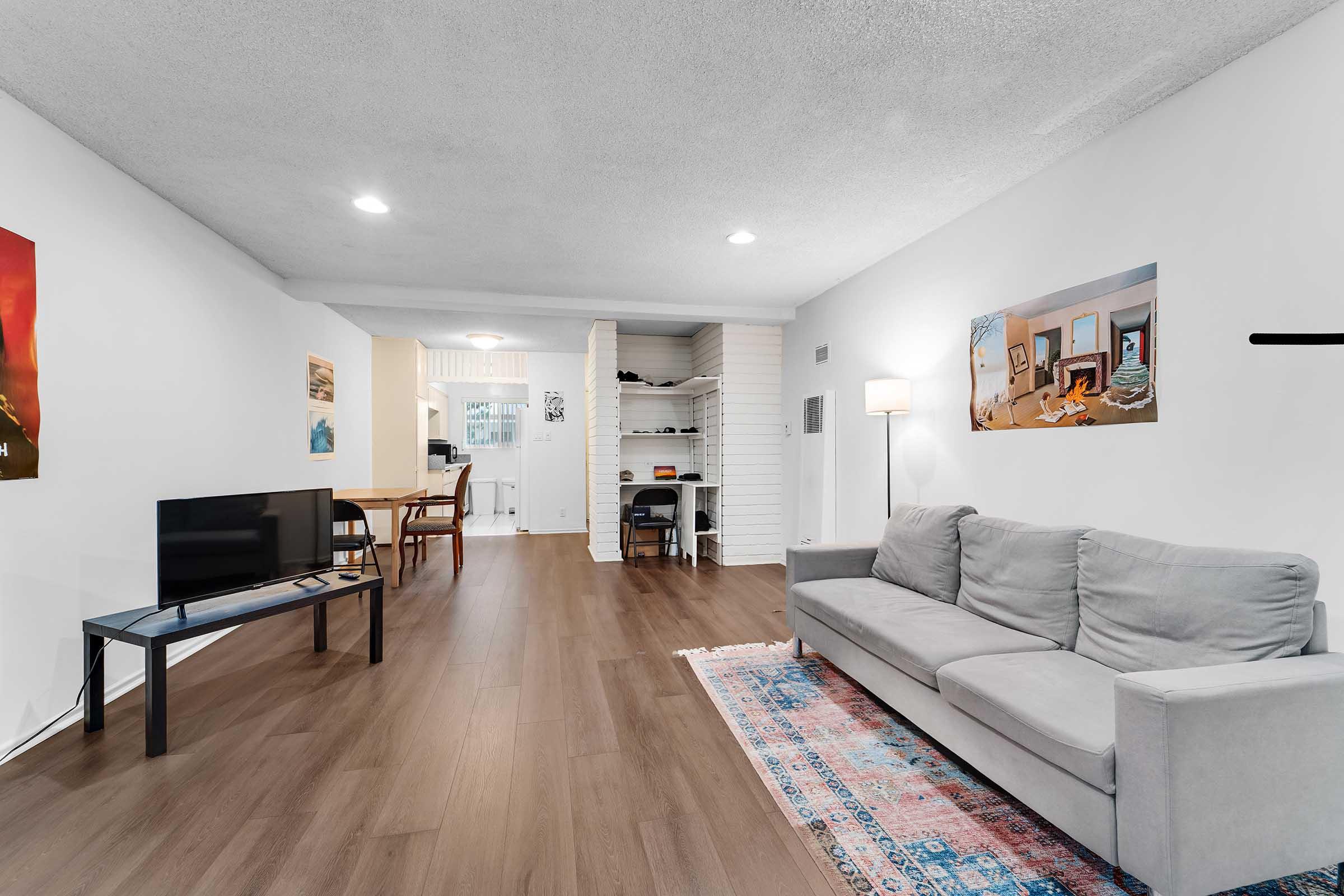 Living room in a modern apartment featuring a gray couch, a television on a black stand, and artwork on the walls. The room has wooden flooring, a light fixture, and a view into an adjacent dining area with a table and shelves. Natural light illuminates the space, creating a cozy ambiance.