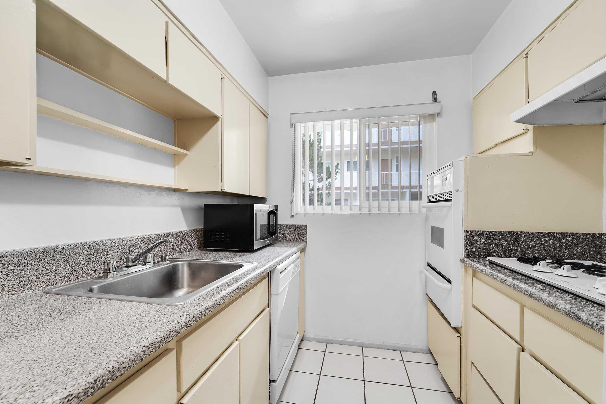 A small kitchen featuring beige cabinetry, a granite countertop, stainless steel sink, microwave, and a white stove. There's a window with blinds allowing natural light, and tiled flooring completes the space. The design is functional and compact, suitable for efficient cooking.