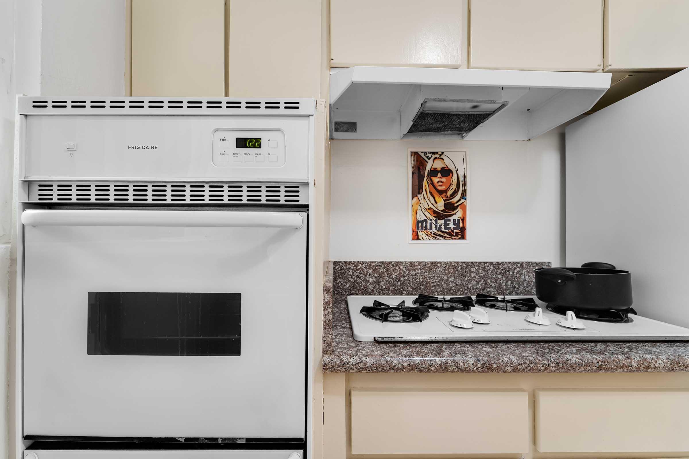 A kitchen with a white refrigerator and an oven. There is a stove with four burners on a granite countertop. A pot sits on the stove, and a framed image of a person in a hooded garment is mounted on the wall above the counter. The overall color scheme is light and neutral.