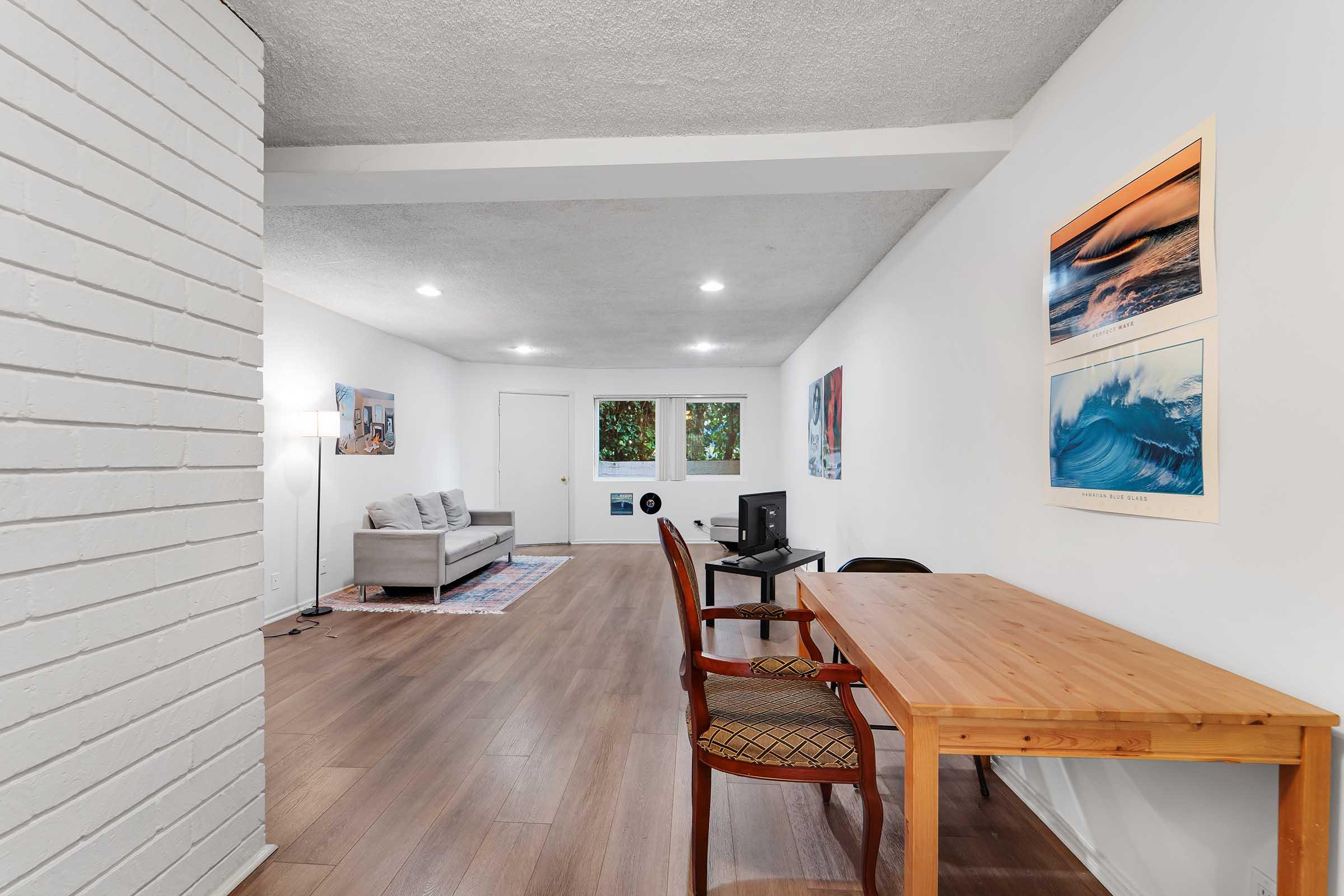 A bright, minimalistic living space featuring a gray sofa, a wooden table with chairs, and artwork on the walls. There are large windows allowing natural light, and a carpet under the sofa. The flooring is wooden, and the walls are painted white, creating an open and airy atmosphere.