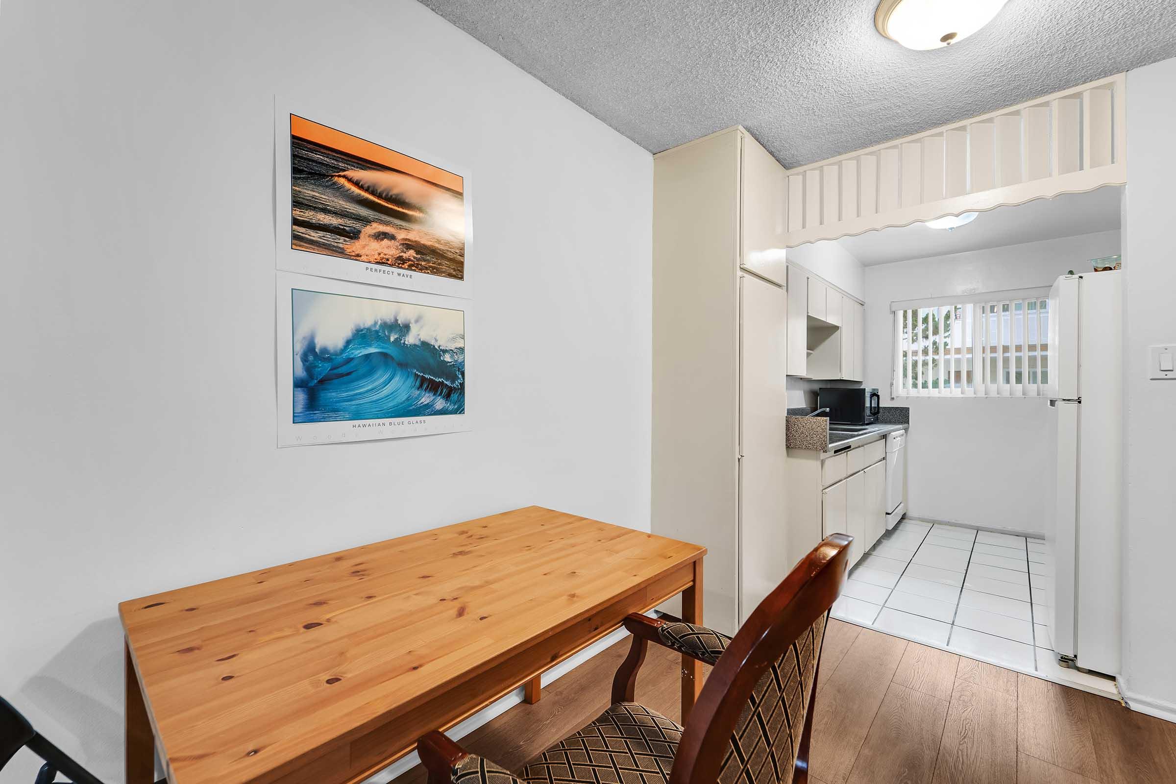 A cozy kitchen area featuring a wooden table and two chairs. The walls are adorned with ocean-themed artwork, and the kitchen includes white cabinets, a refrigerator, and a window providing natural light. The floor is tiled, complementing the overall clean and inviting atmosphere.