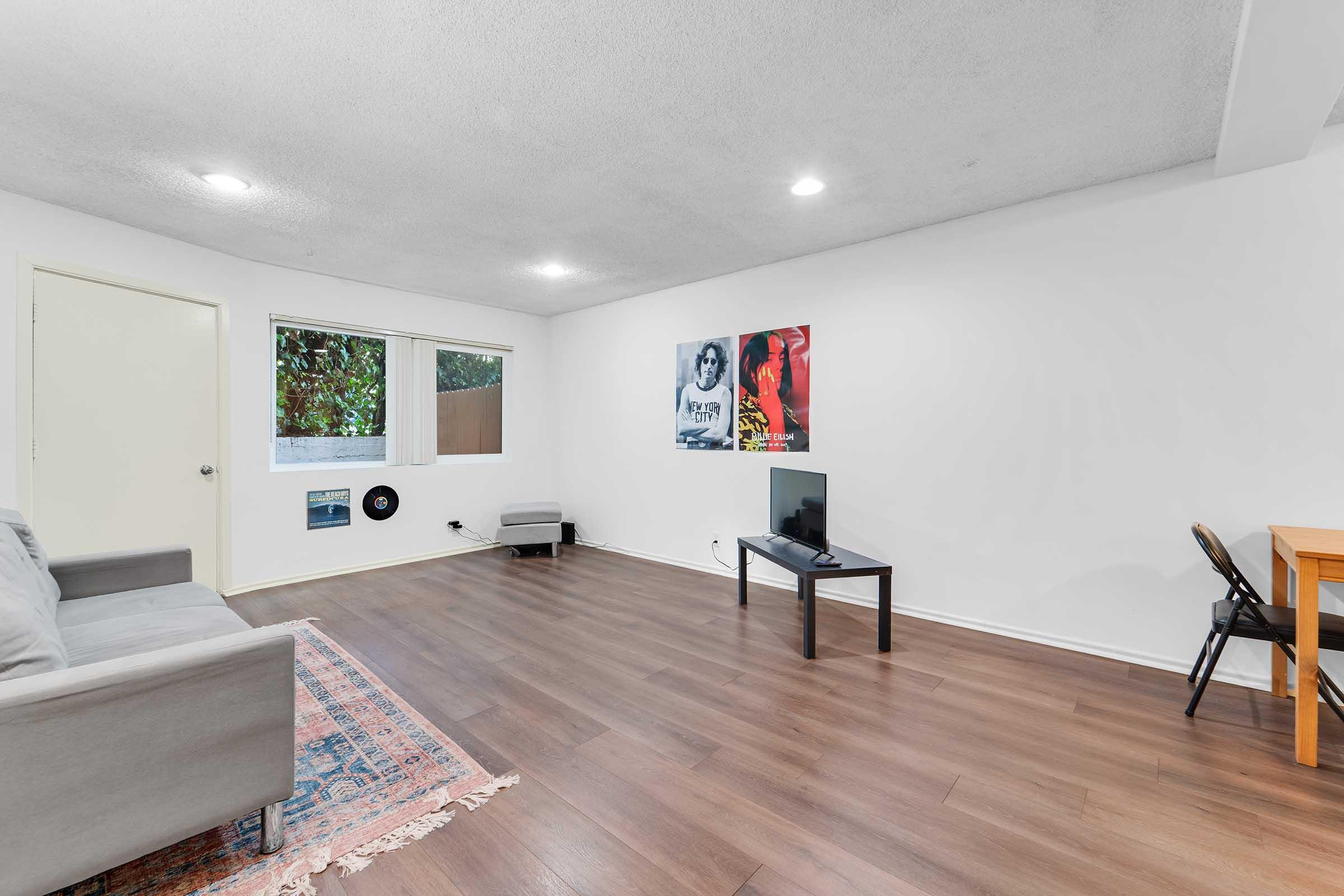 A living room with light-colored walls and wooden flooring. There is a gray sofa, a small chair, a television stand with a TV, and a dining table with a chair. On one wall, there are two framed artworks. Large windows provide natural light, and a patterned rug is on the floor.