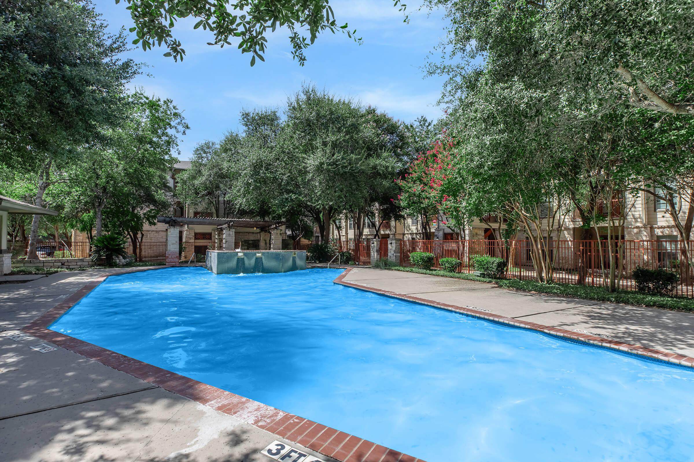 A sunny outdoor swimming pool surrounded by lush green trees and landscaped bushes. The pool features a small fountain on one side and is bordered by a concrete deck. In the background, there are apartment buildings visible, adding to a peaceful residential atmosphere.