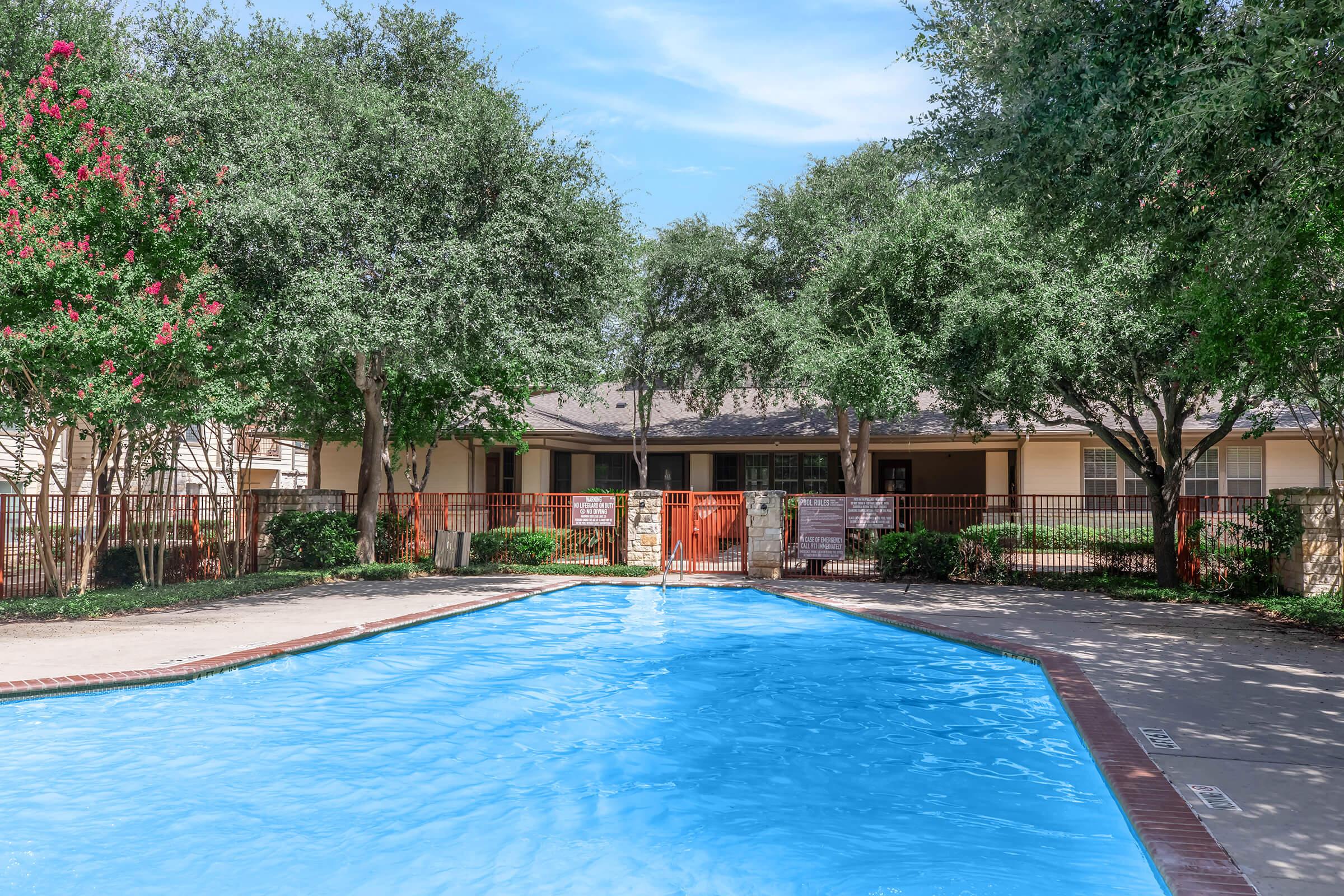 A clear blue swimming pool surrounded by lush greenery and trees, with a fence and a building in the background. The scene is bright and inviting, featuring a sunny sky and well-maintained landscaping, ideal for relaxation.