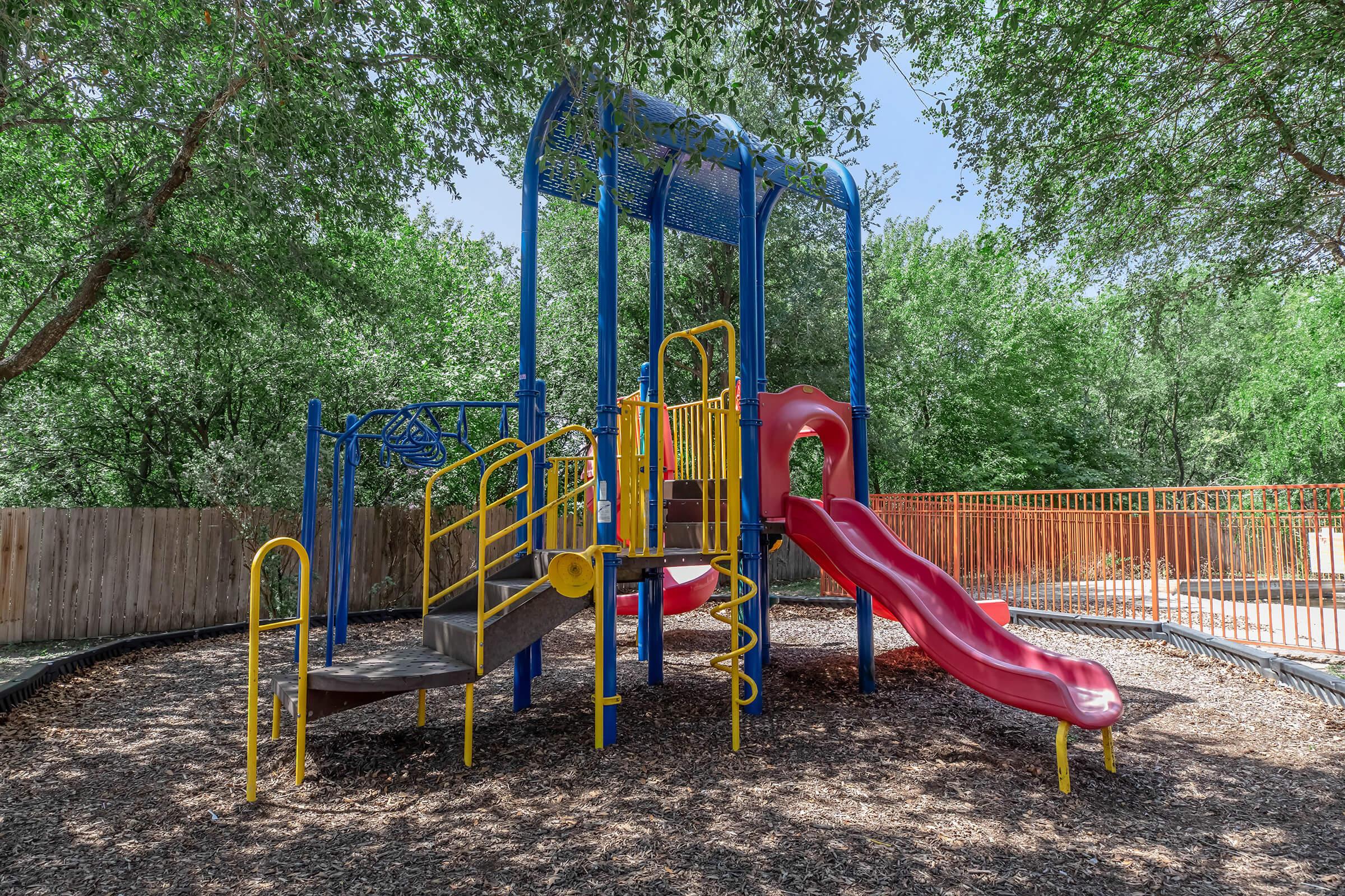 Colorful playground structure featuring a red slide, climbing features, and yellow railings, surrounded by lush green trees. The ground is covered in wood chips, and there is a wooden fence in the background.