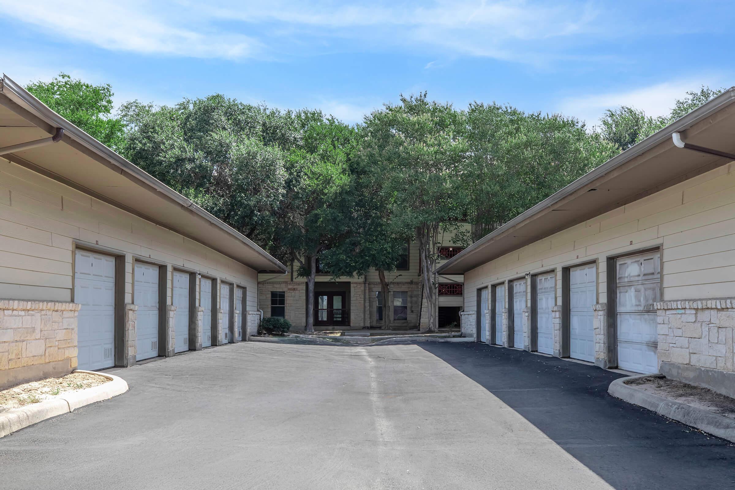 A view of a wide, paved driveway leading to two rows of garage doors with a backdrop of green trees and a building. The scene is sunny and calm, showcasing a residential area with ample parking space and well-maintained surroundings.