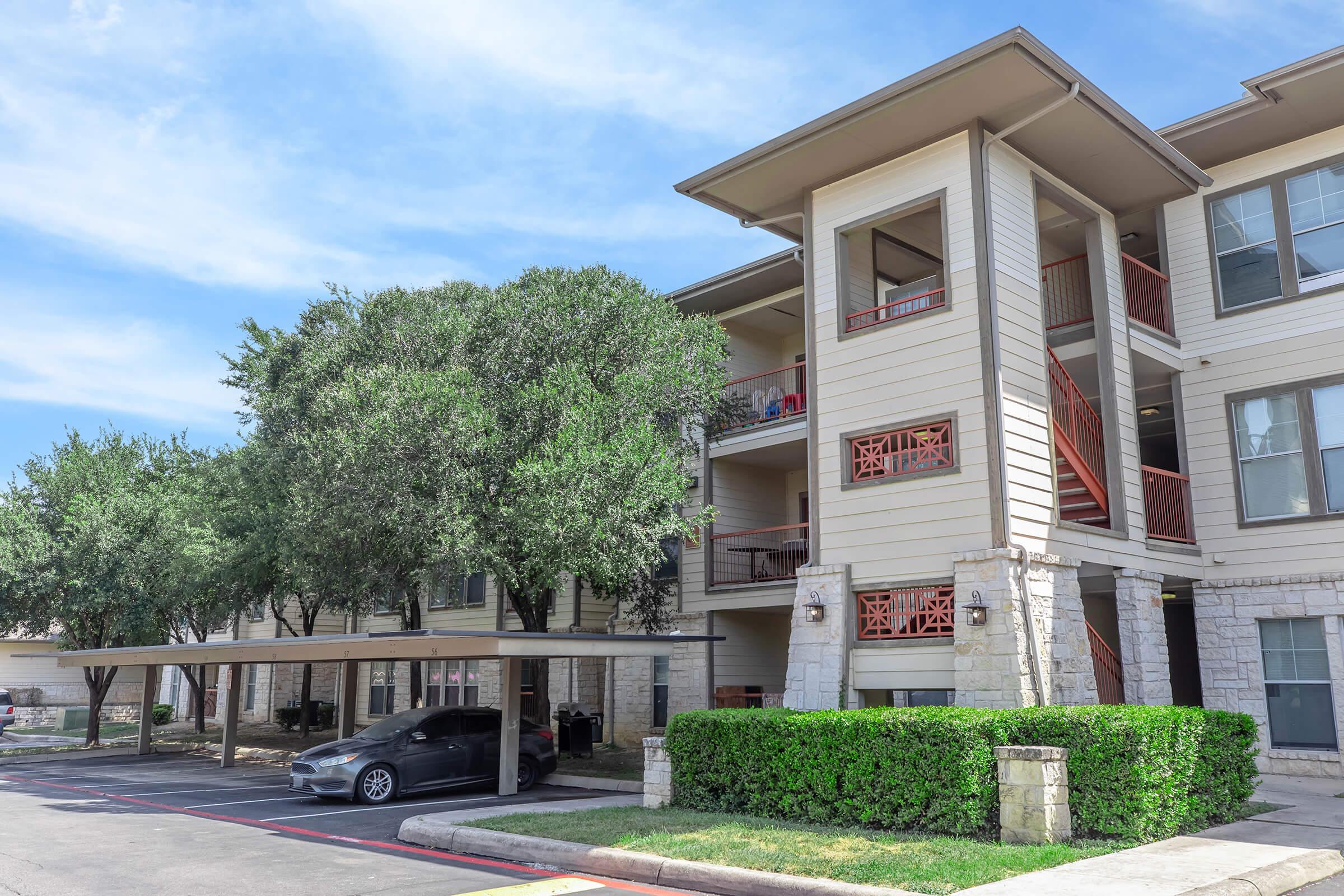 Three-story apartment building with a modern design, featuring beige siding and red accents. There are shaded parking spaces in front and well-maintained greenery including bushes and trees. The sky is clear with some clouds.