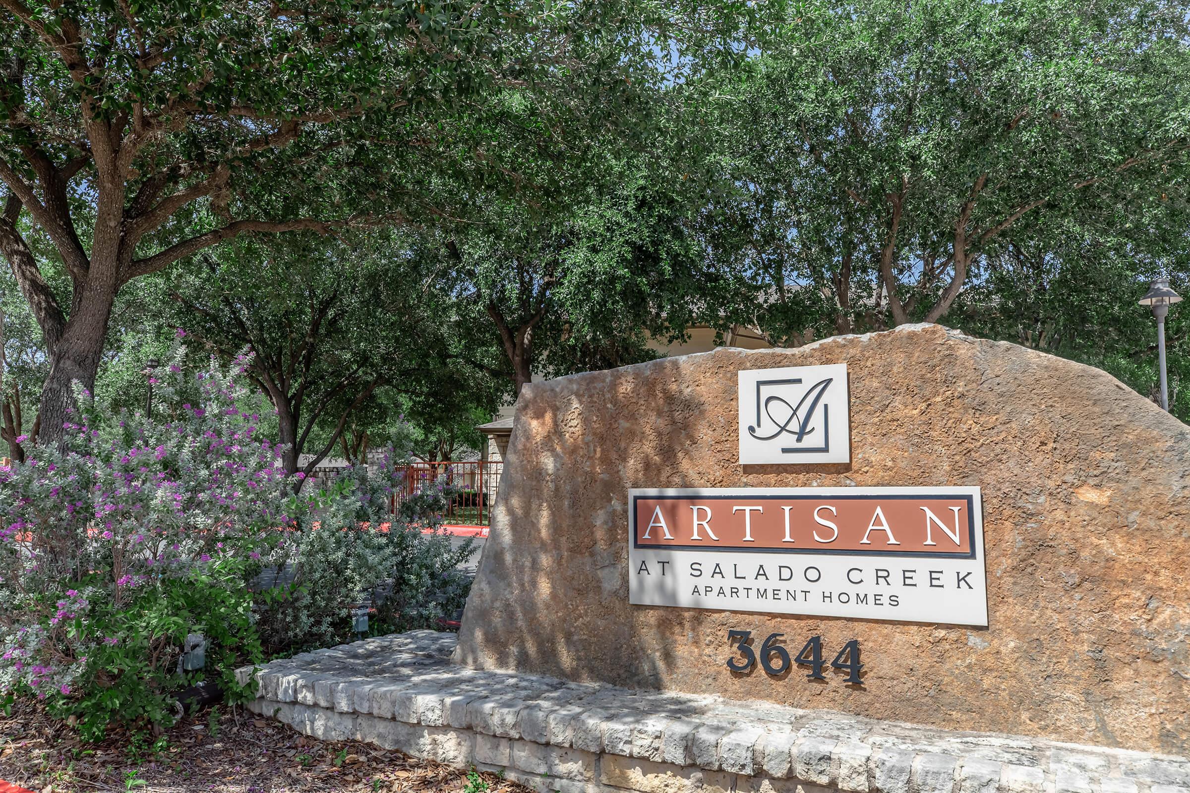 Sign for Artisan at Salado Creek Apartment Homes, featuring the name 'ARTISAN' prominently displayed on a large, natural stone. Surrounding the sign are lush green trees and colorful flowering shrubs, creating an inviting atmosphere. The street number '3644' is also included on the sign.