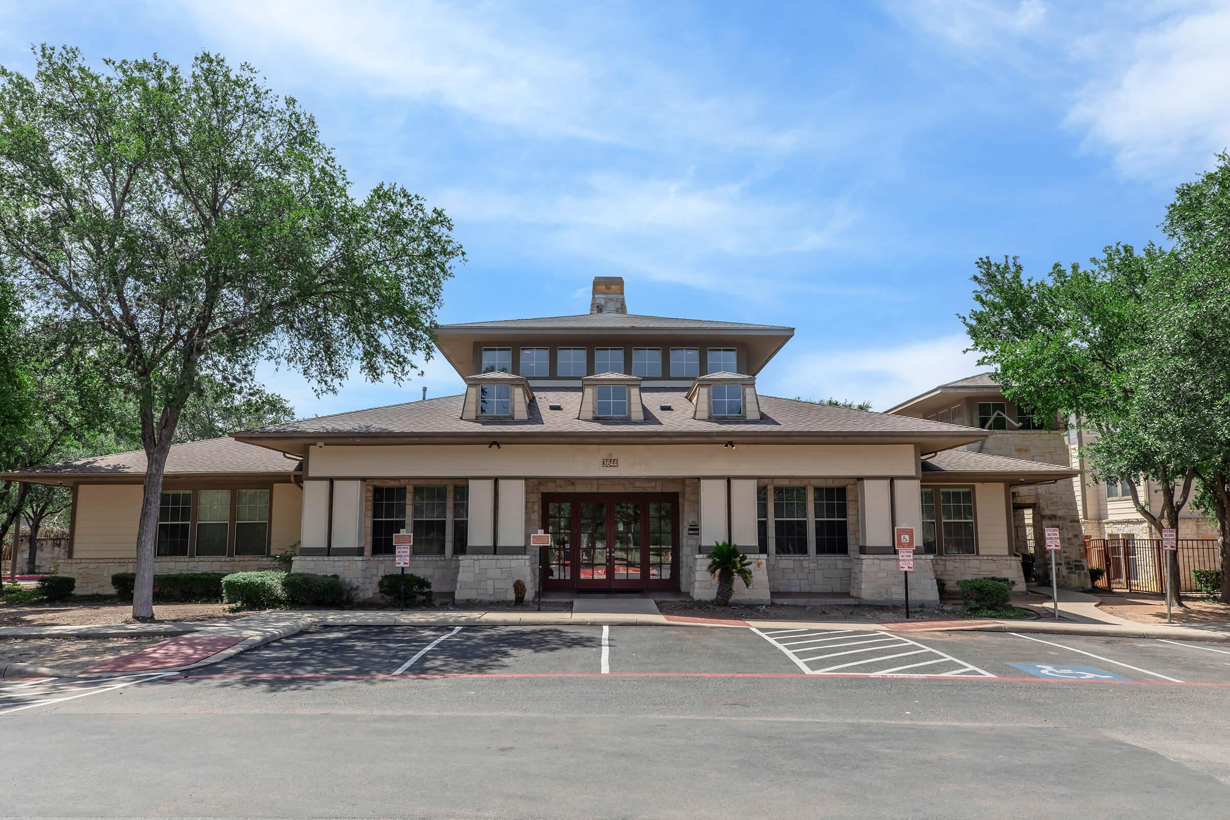 A two-story building with a sloped roof and large front windows, surrounded by trees and parking spaces. The entrance features double doors and is flanked by shrubs. The sky is clear with a few clouds, indicating a sunny day.