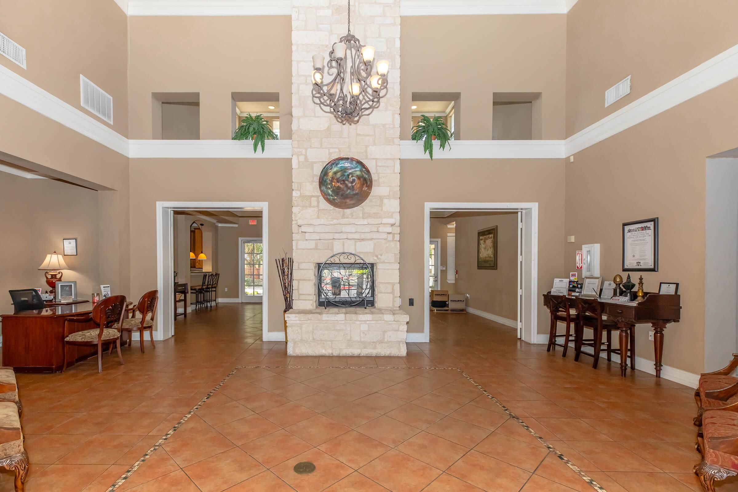 Spacious lobby with warm, neutral walls and tiled flooring. A large stone fireplace is the focal point, flanked by potted plants. Bright light fills the room from large windows. Elegant furniture, including chairs and a desk, is arranged in inviting areas. A chandelier hangs from the ceiling, adding a touch of sophistication.