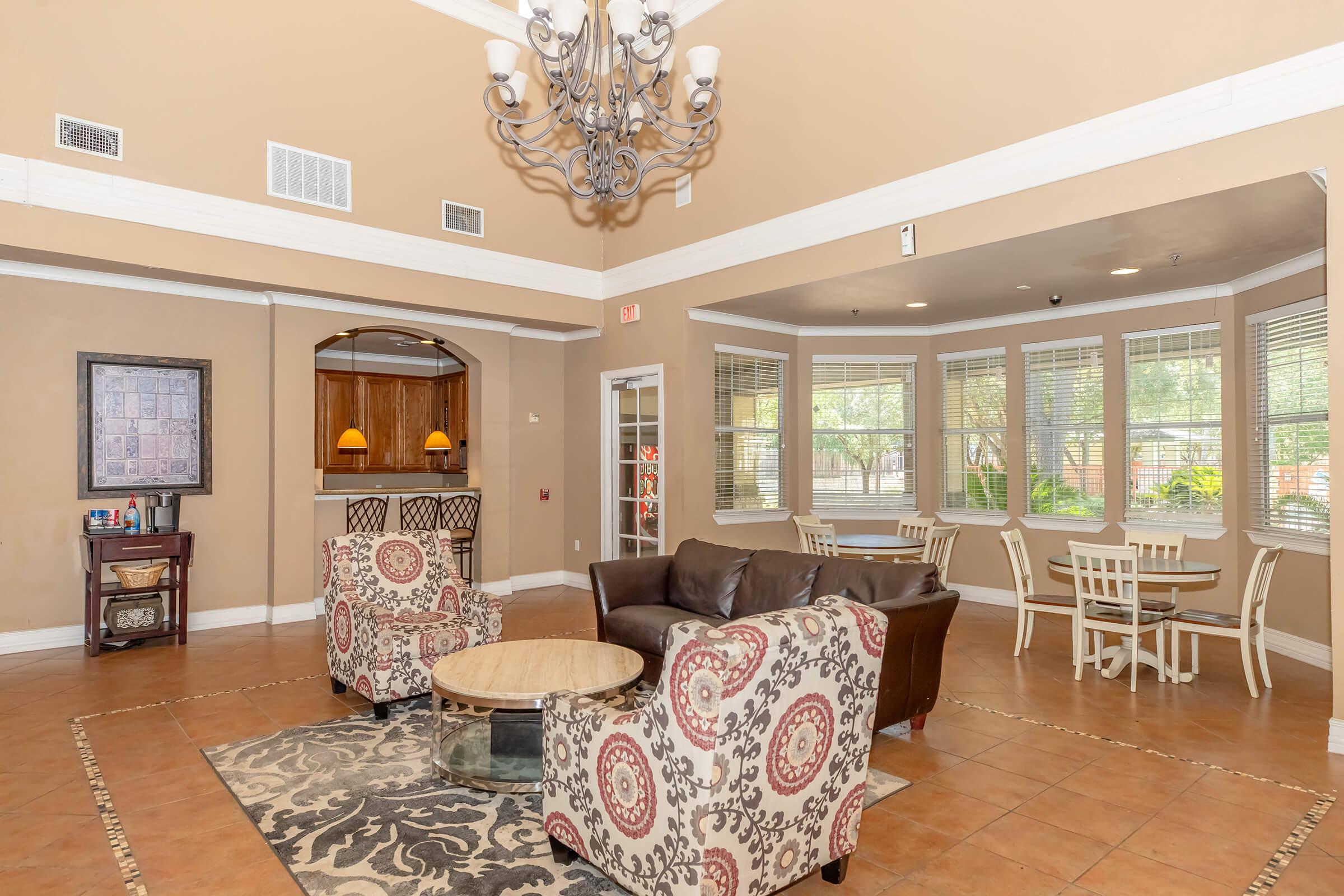 A cozy community lounge area featuring a large chandelier, comfortable seating with patterned chairs, a central wooden coffee table, and small dining tables near windows. The walls are painted in warm tones, creating an inviting atmosphere with natural light streaming in.