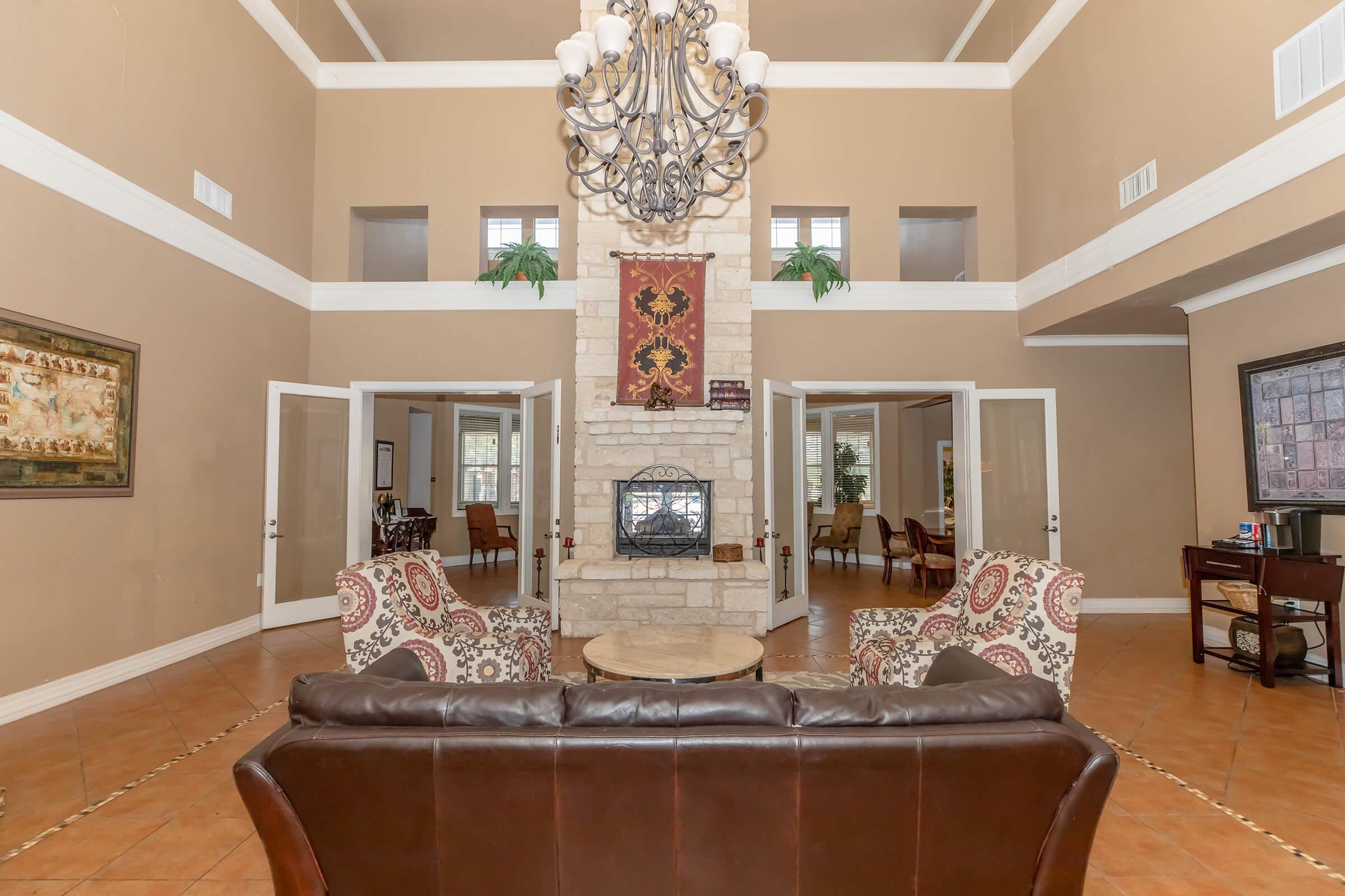 A spacious living area featuring high ceilings, a stone fireplace, and a chandelier. Two patterned armchairs are placed around a central table, with large windows providing natural light. The walls are painted a warm tan, and decorative plants and artwork add character to the room.