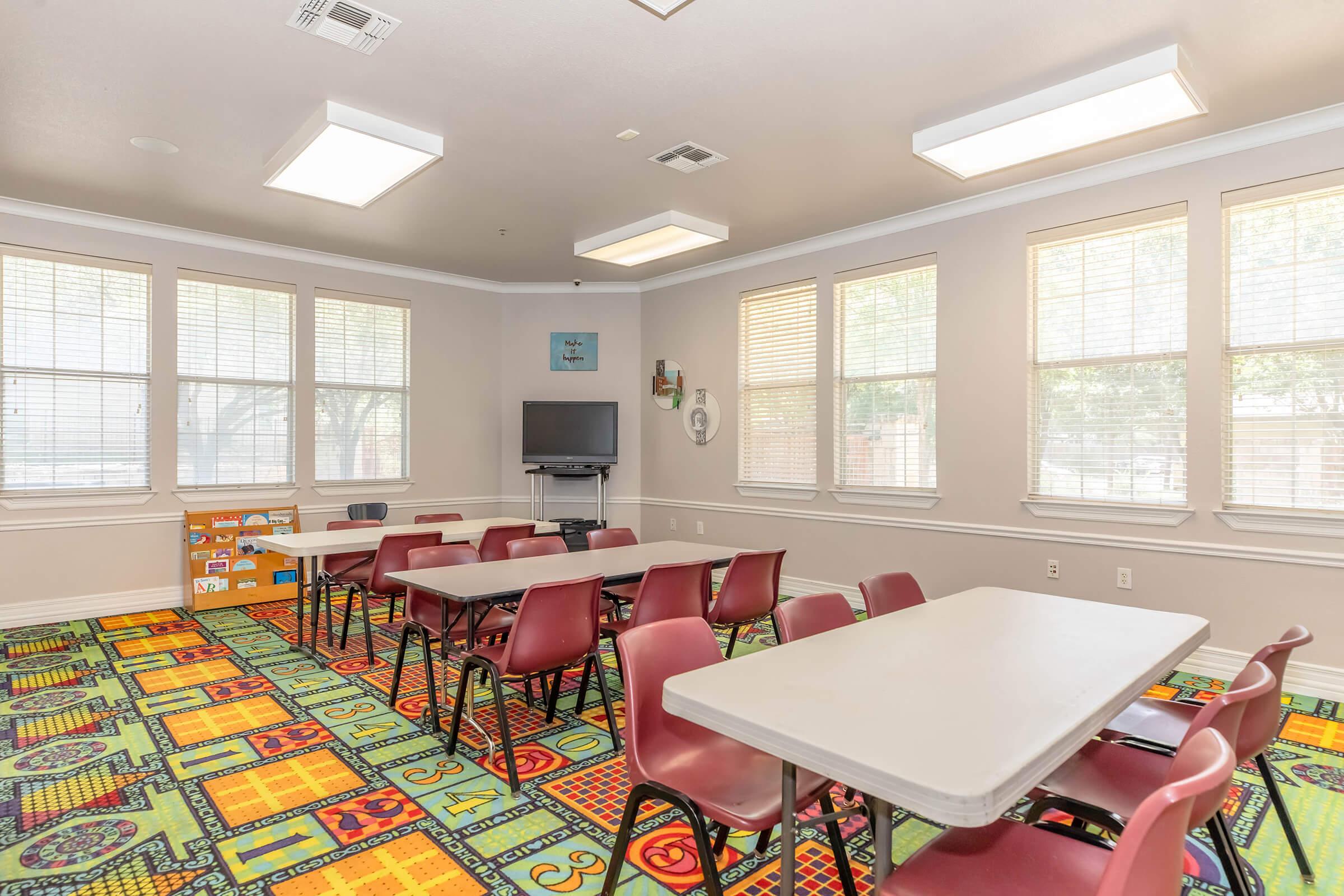 A bright classroom or activity room with several tables and chairs arranged for use. The floor features colorful, patterned carpeting, and there are large windows allowing natural light to fill the space. A television is mounted on the wall, with artwork and a clock decorating the room.