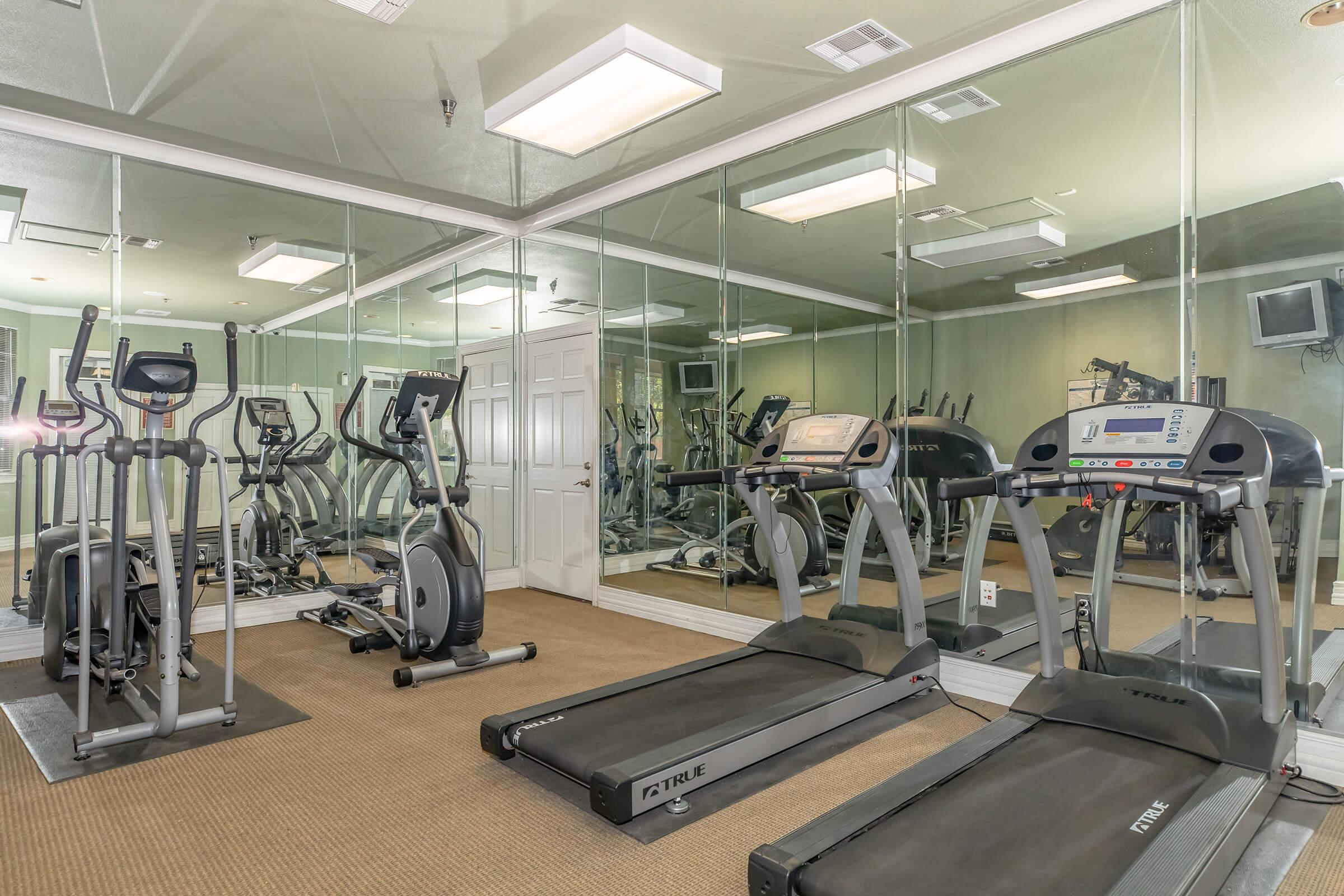 A clean gym area featuring two treadmills and an elliptical machine, all facing a large mirror. The room has soft lighting and a neutral carpet, with additional workout equipment visible in the background. A flat-screen TV is mounted on the wall.