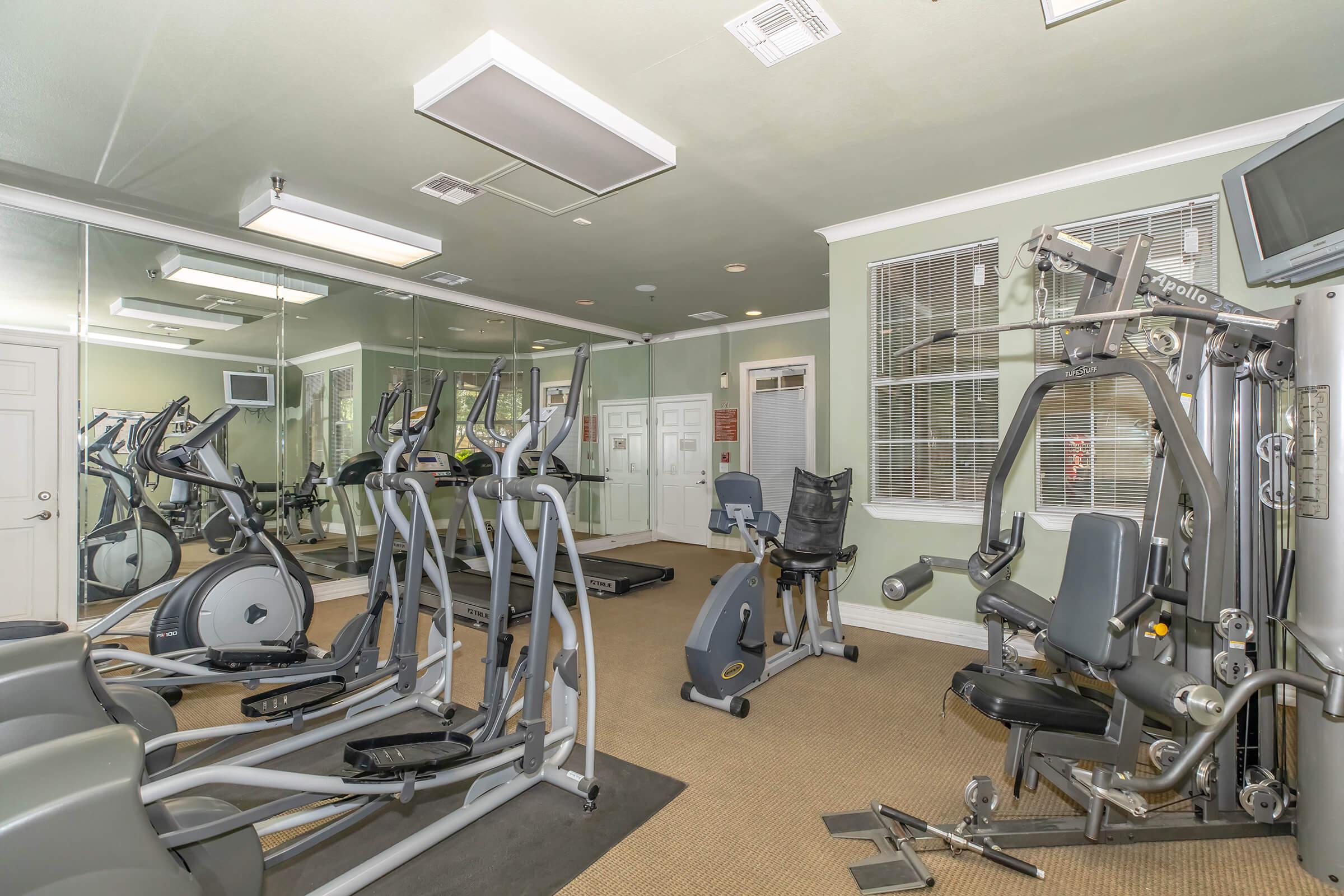 A modern gym with various fitness equipment, including treadmills, ellipticals, and a multi-station machine. The room features mirrors, soft overhead lighting, and neutral-colored walls, creating a spacious and inviting workout environment. Large windows let in natural light.
