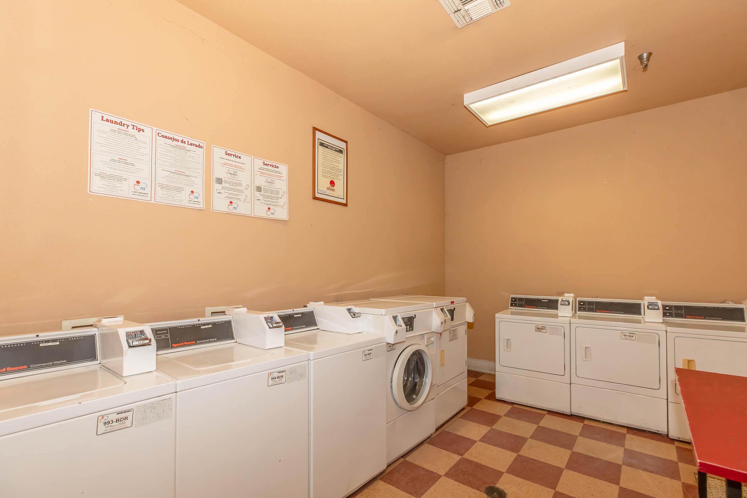 A laundry room featuring several white washing machines and dryers along one wall, with a light beige wall in the background. The floor has a checkered pattern of light and dark beige tiles. There are informational posters hanging on the wall above the machines.
