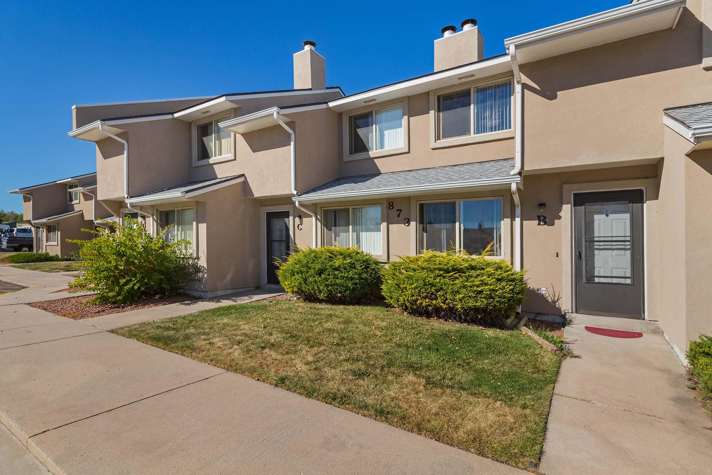 Row of townhouses with a light beige exterior, featuring green shrubs in front. Each unit is labeled with numbers and letters. A clear blue sky is visible above, enhancing the bright atmosphere of the neighborhood. Concrete walkways lead to the doorsteps of each unit.