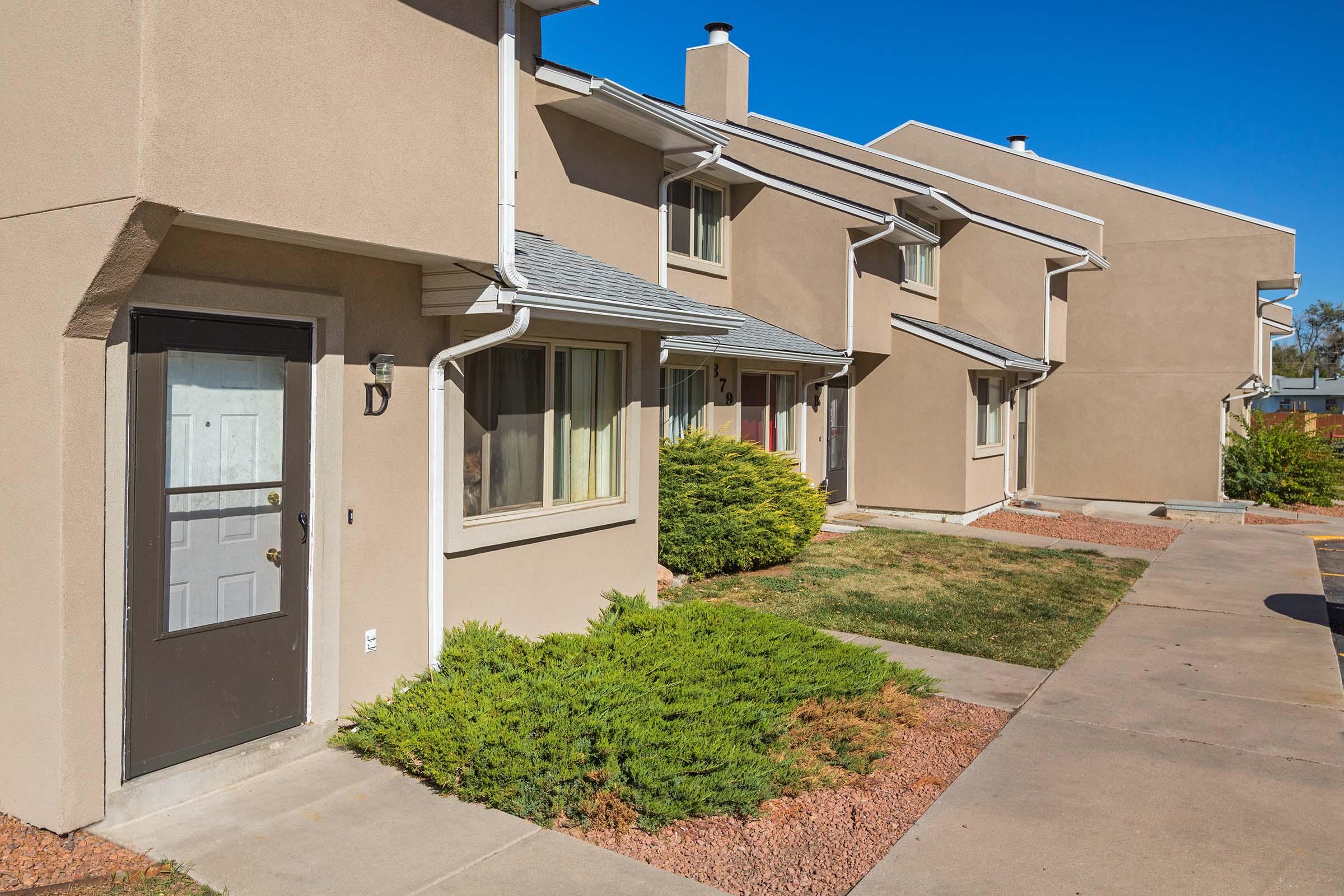 Row of residential buildings featuring beige exteriors and small front gardens with shrubs. Each unit has a door, windows, and a walkway, set against a clear blue sky. The area appears well-maintained, suggesting a suburban living environment.