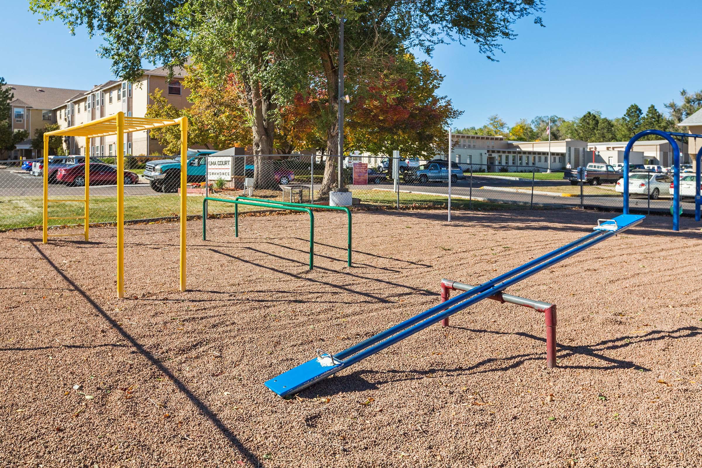 A playground area featuring a yellow climbing frame, green parallel bars, and a blue seesaw on gravel. In the background, there are residential buildings and trees with autumn foliage under a clear blue sky.