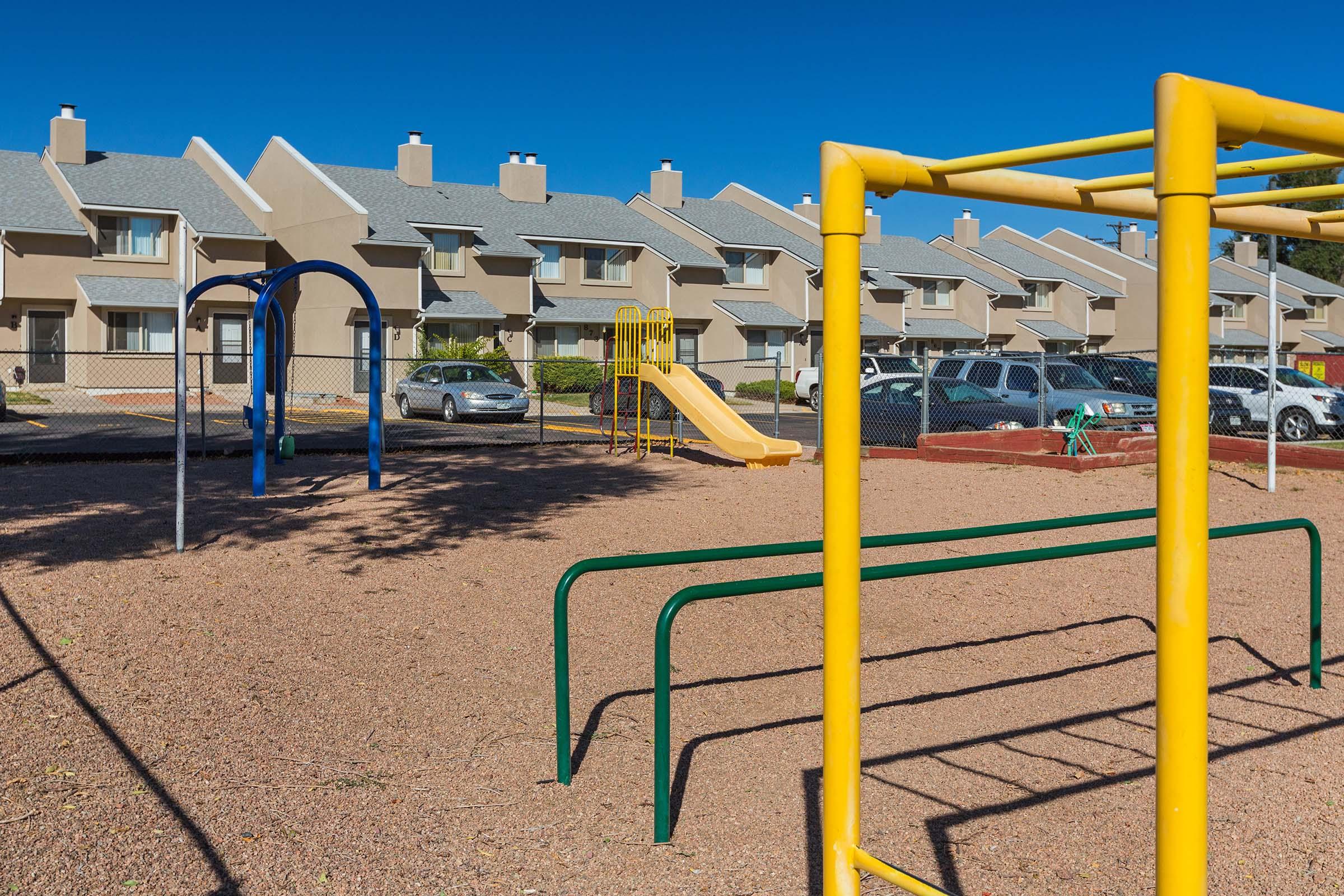 A playground featuring a blue swing set, a yellow slide, and green climbing bars, with a sandy ground. In the background, there are rows of residential buildings with a clear blue sky above. Several parked cars are visible behind the playground area.
