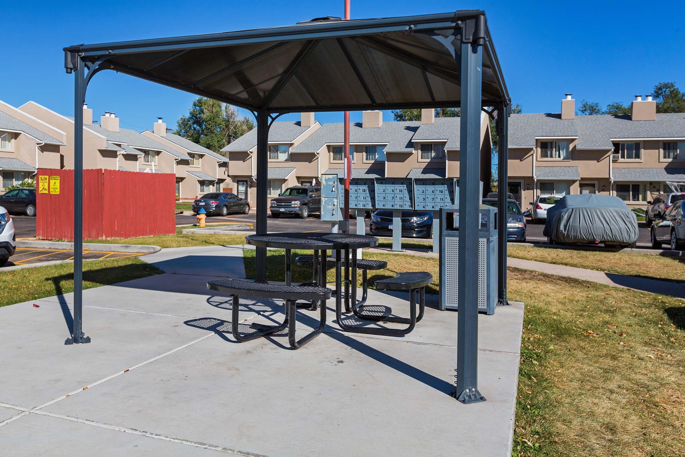 A shaded picnic area with a metal table and benches under a gazebo, surrounded by a grassy space. In the background, several residential buildings and parked cars are visible. Mailboxes are situated on one side of the picnic area. Bright blue sky overhead indicates a clear day.