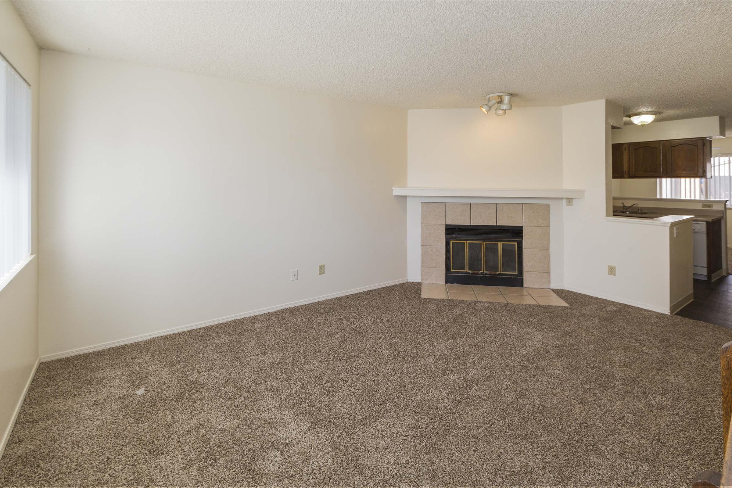 Spacious living room featuring light-colored walls, a carpeted floor, and a fireplace with a tiled area. A doorway leads to a kitchen visible in the background, with cabinets and appliances. Large windows allow natural light to fill the room.