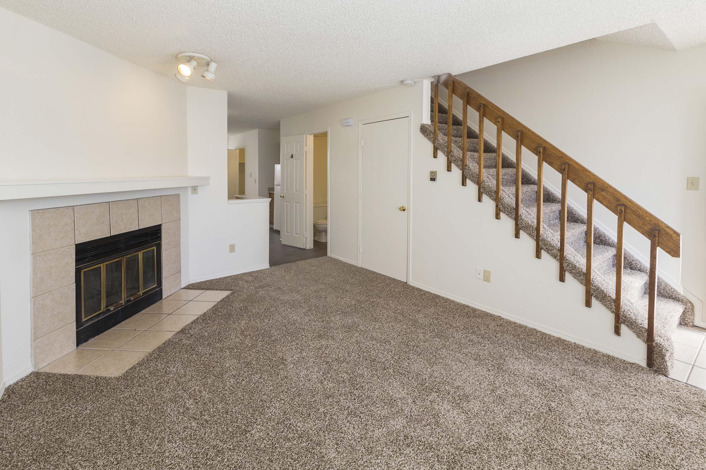 A spacious living area featuring a carpeted floor, a fireplace with a tile surround, and a wooden staircase leading to the upper level. Light-colored walls and natural light create a bright atmosphere. The room is designed for comfort and cozy living.