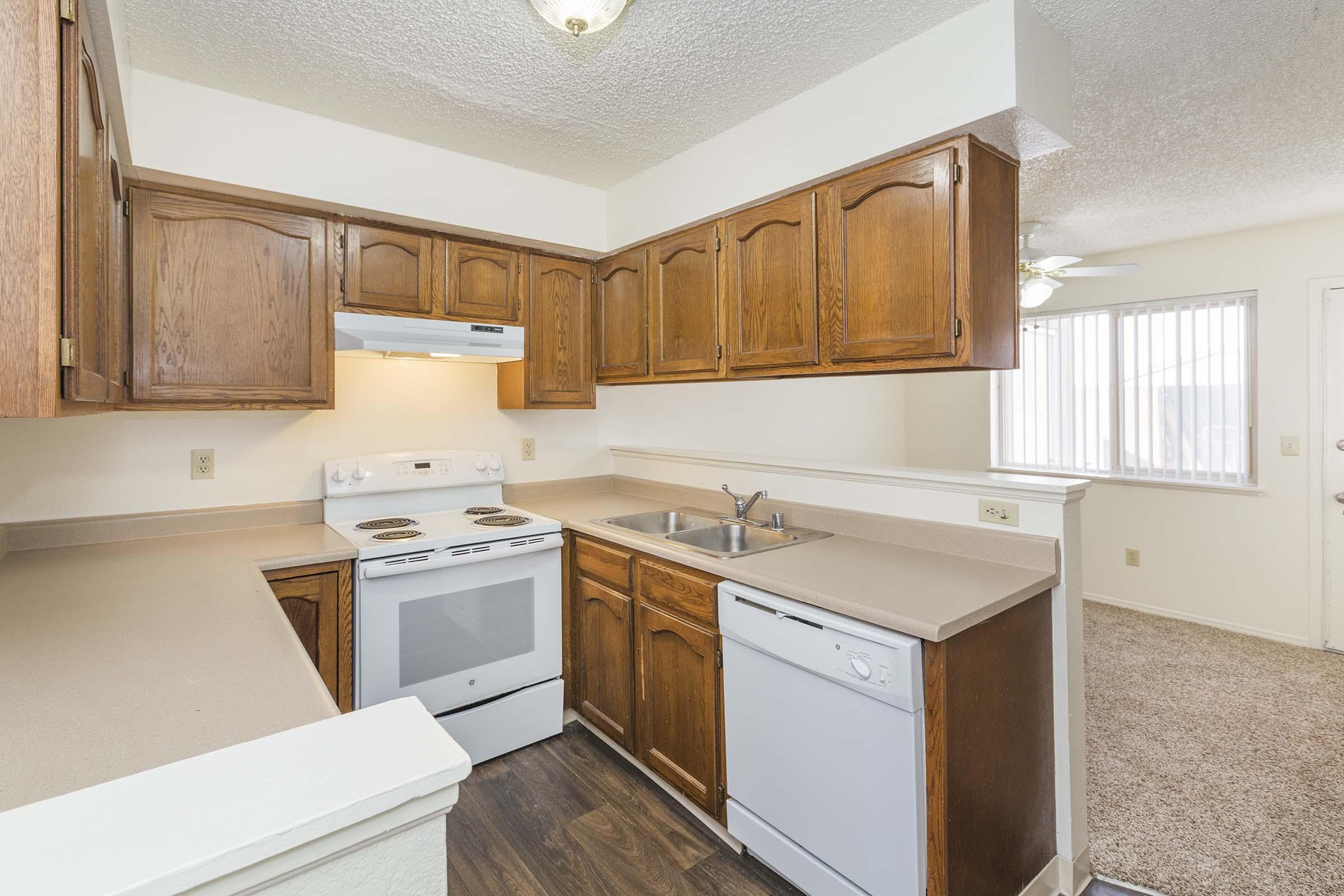 A small, well-lit kitchen featuring wooden cabinets, a white stove, and a dishwasher. The countertop is beige, and there's a sink with a faucet. Natural light streams in through a window. The adjoining space has beige carpeting and a door leading outside.