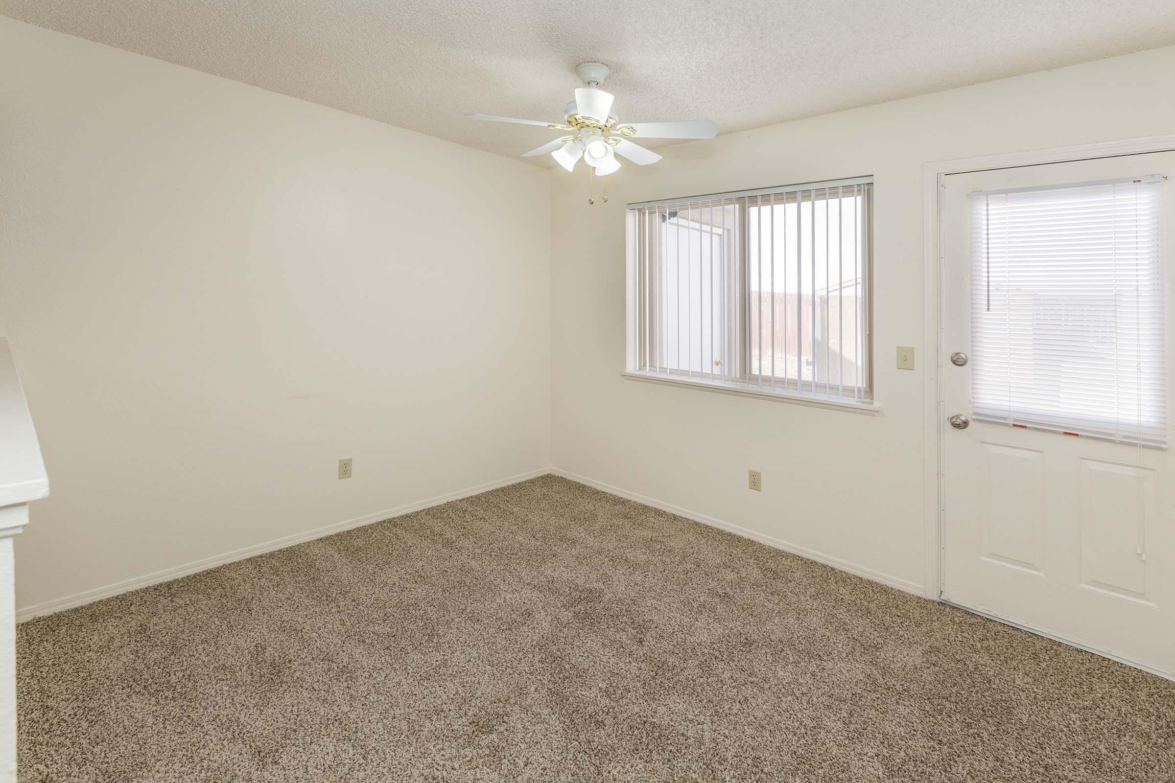 An empty room with light-colored walls and plush carpeting. It features a ceiling fan, a large window with blinds, and a door leading outside. The space is well-lit and clean, creating a cozy, inviting atmosphere.