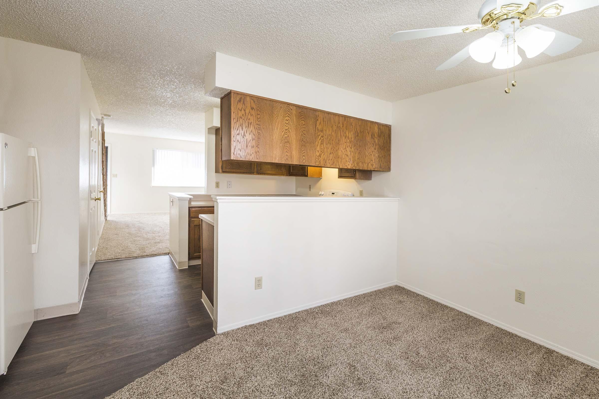 A bright, open-plan kitchen and living area featuring a ceiling fan, wooden cabinetry, and a light-colored wall. The kitchen has a refrigerator and a glimpse of carpeted flooring in the adjoining living space, creating a cozy atmosphere.