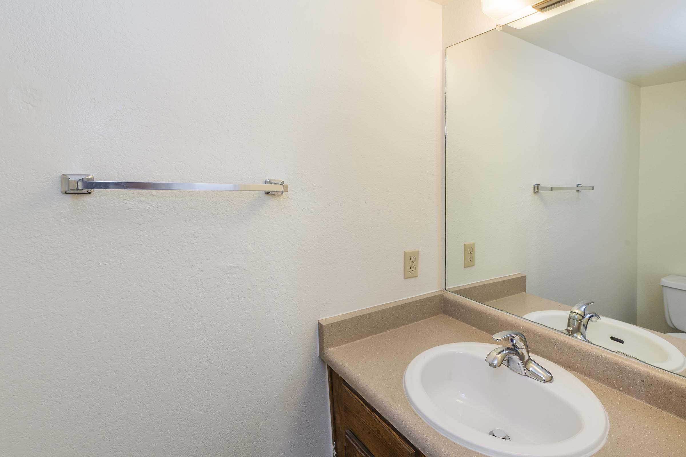 A clean bathroom with a light-colored wall, a small sink with a chrome faucet and countertop, a large mirror, and a towel bar. There is a light fixture above the mirror and a toilet partially visible to the right. The overall atmosphere is simple and modern.
