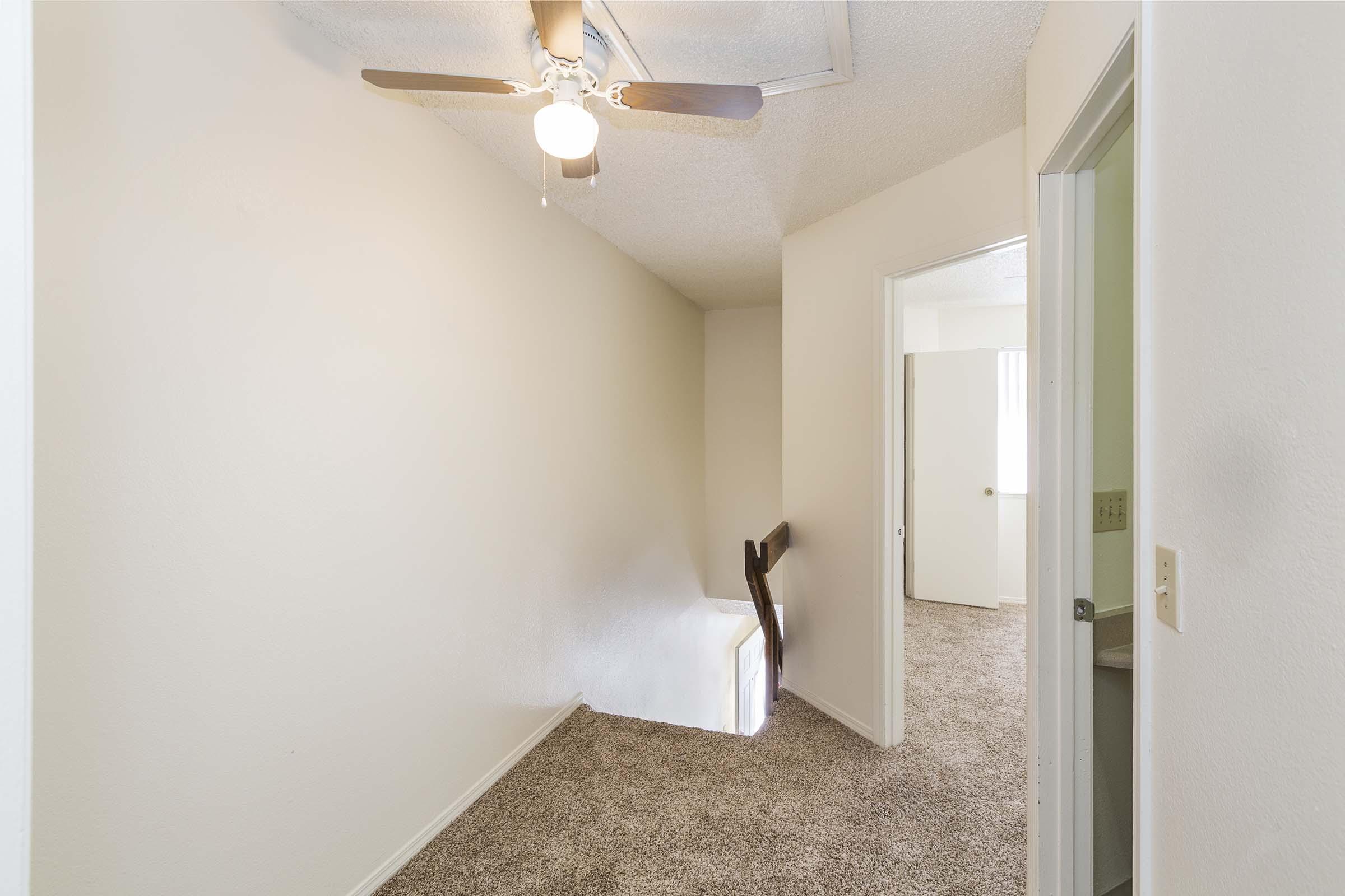 A well-lit hallway featuring beige carpet, a ceiling fan, and white walls. A staircase is visible on the left side, leading down, while a door is seen in the background, indicating access to another room. The overall atmosphere is airy and inviting.
