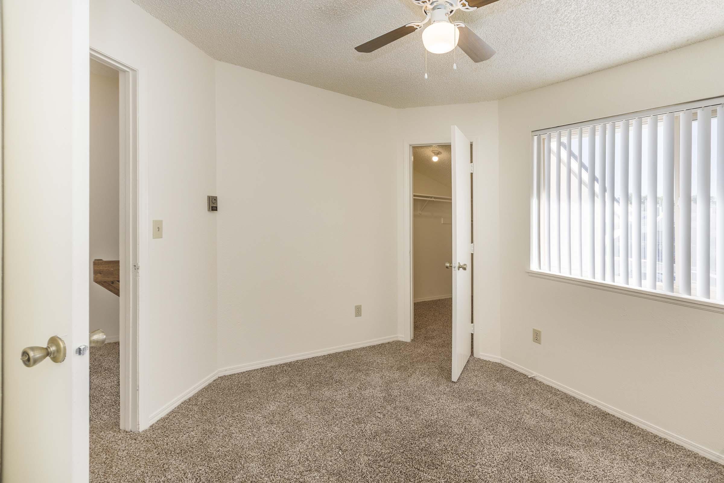 A well-lit room with beige carpet and white walls. It features a ceiling fan, a window with vertical blinds, and an open door leading to a closet. Another door is visible on the left, suggesting access to a hallway or another room. The space appears clean and inviting.