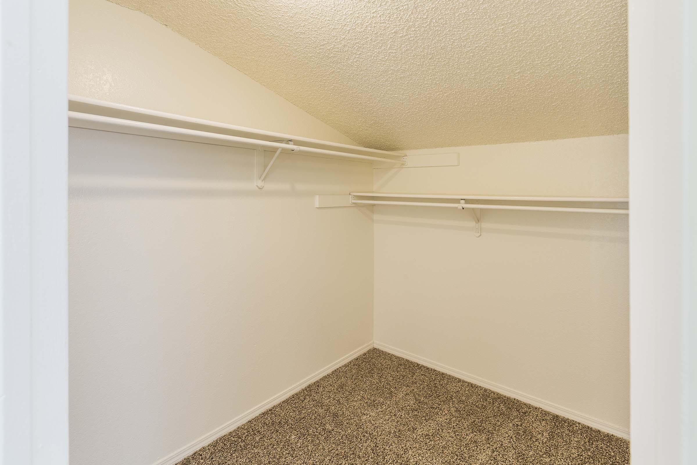 Empty closet with slanted ceiling, featuring white shelving and carpeted flooring. Walls are painted in a light color, creating a bright and spacious appearance.