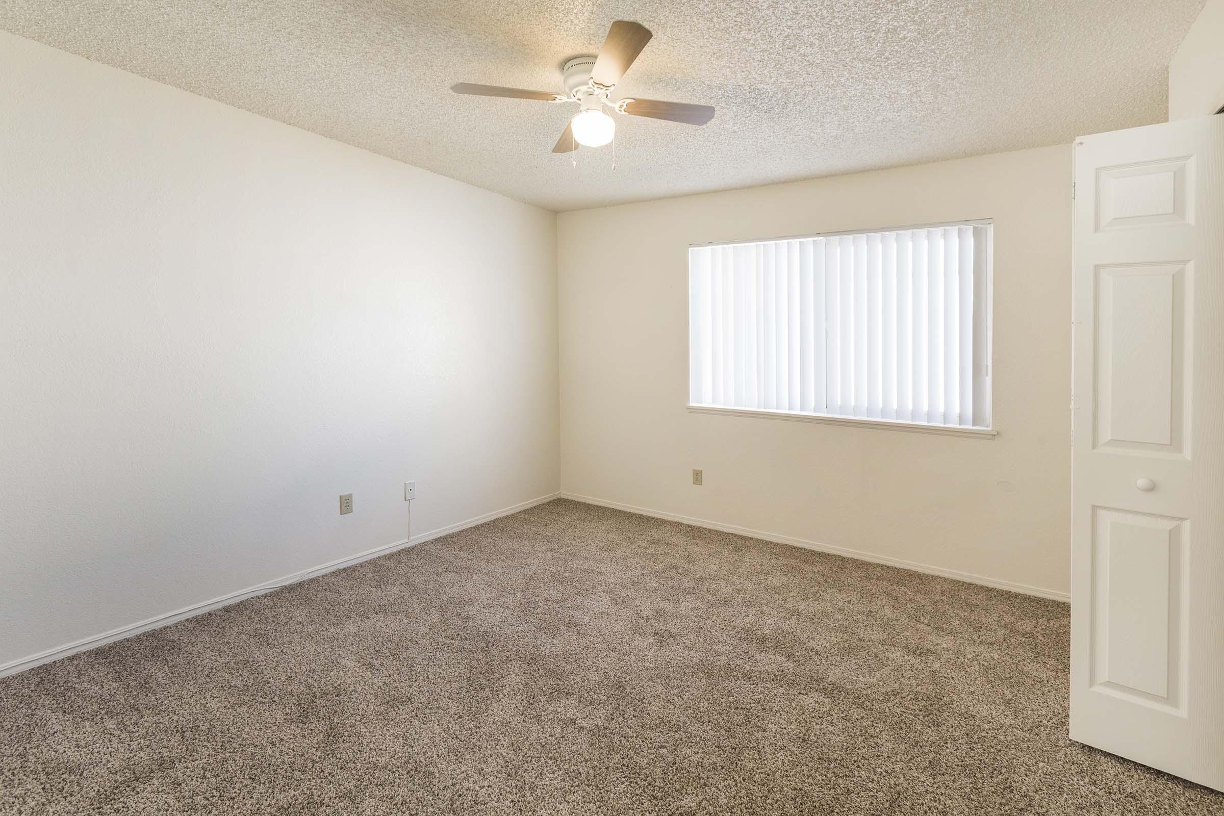 A bright, empty room with light-colored walls and a ceiling fan. The floor is covered in beige carpet, and there is a window with vertical blinds on one wall. A closed white door is visible on the right side, suggesting access to another space. The overall atmosphere is clean and inviting.