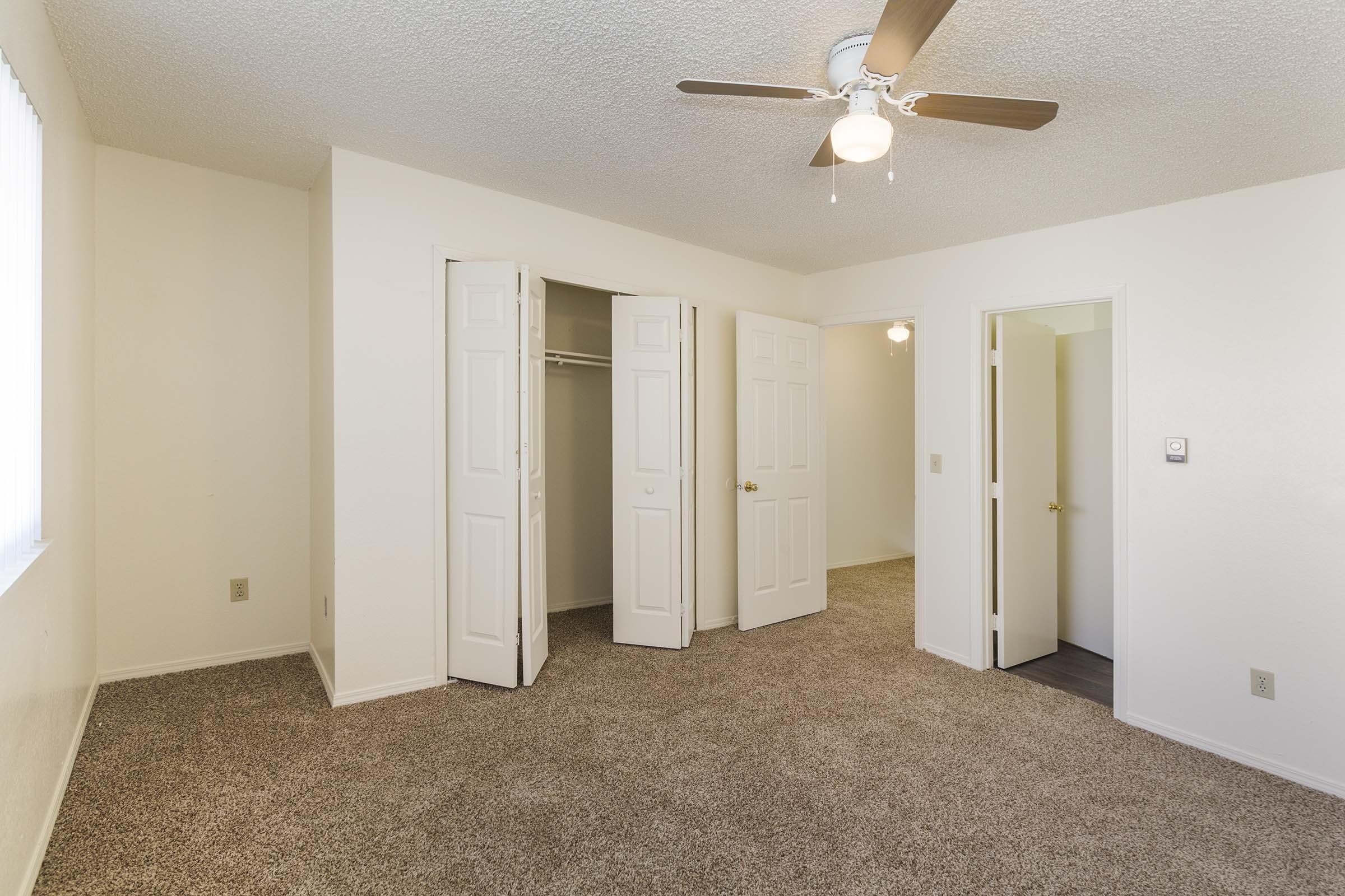 Spacious, empty bedroom featuring beige carpet, light-colored walls, a ceiling fan with wooden blades, and two open closet doors. Natural light streams in from a window, illuminating the room's neutral decor. A door leads to an adjoining area, providing a warm and inviting atmosphere.