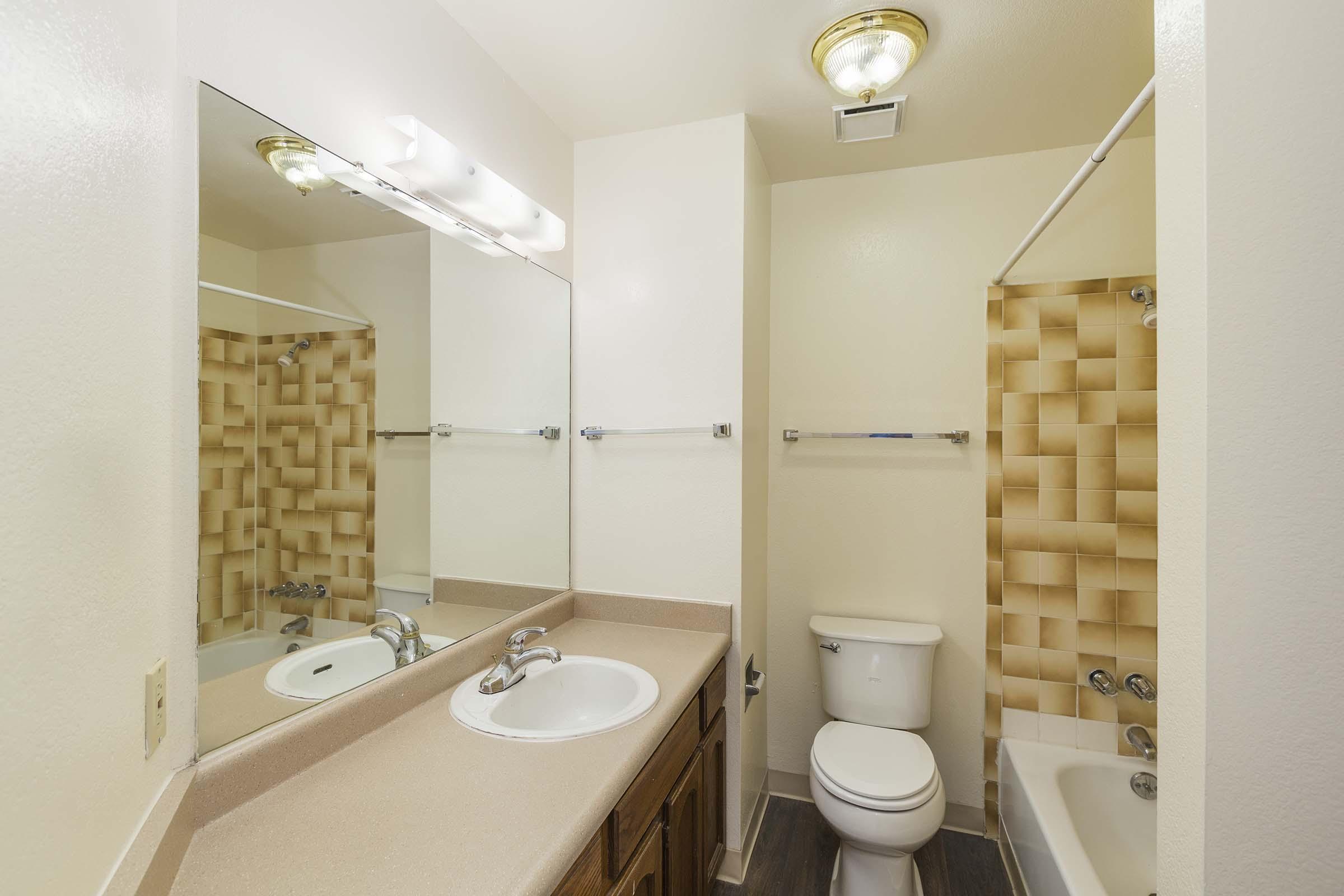 A small bathroom featuring a light-colored countertop with a sink, a wall-mounted mirror, and a flush toilet. The shower area has beige tiled walls, and there is a bathtub with a showerhead. The flooring is dark, contrasting with the lighter walls and fixtures. Simple lighting overhead provides illumination.