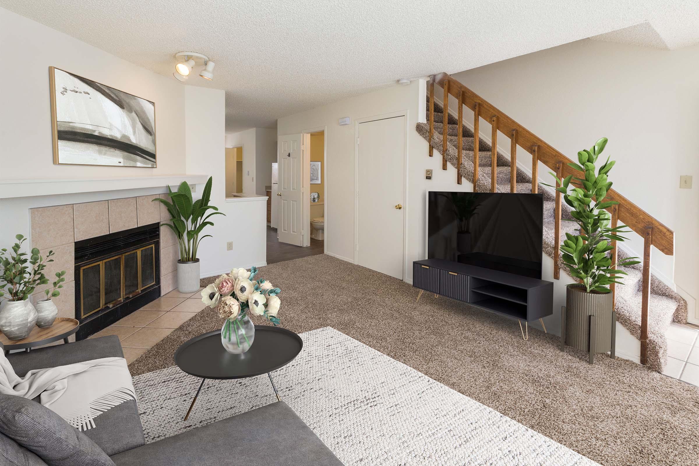A modern living room featuring a cozy seating area with a gray couch, a round black coffee table, and a decorative floral arrangement. The space has a fireplace, a large flat-screen TV on a media stand, and potted plants. A staircase leads to the upper level, with light walls and natural lighting enhancing the inviting atmosphere.