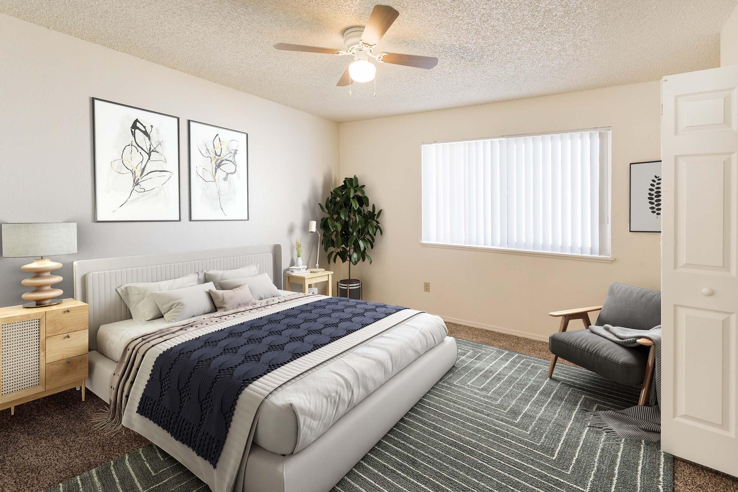 A cozy, well-lit bedroom featuring a large bed with a gray blanket, two decorative pillows, and artwork on the wall. A nightstand with a lamp sits beside the bed, and there is a tall plant in the corner. A comfortable armchair and a window with vertical blinds complete the inviting space.